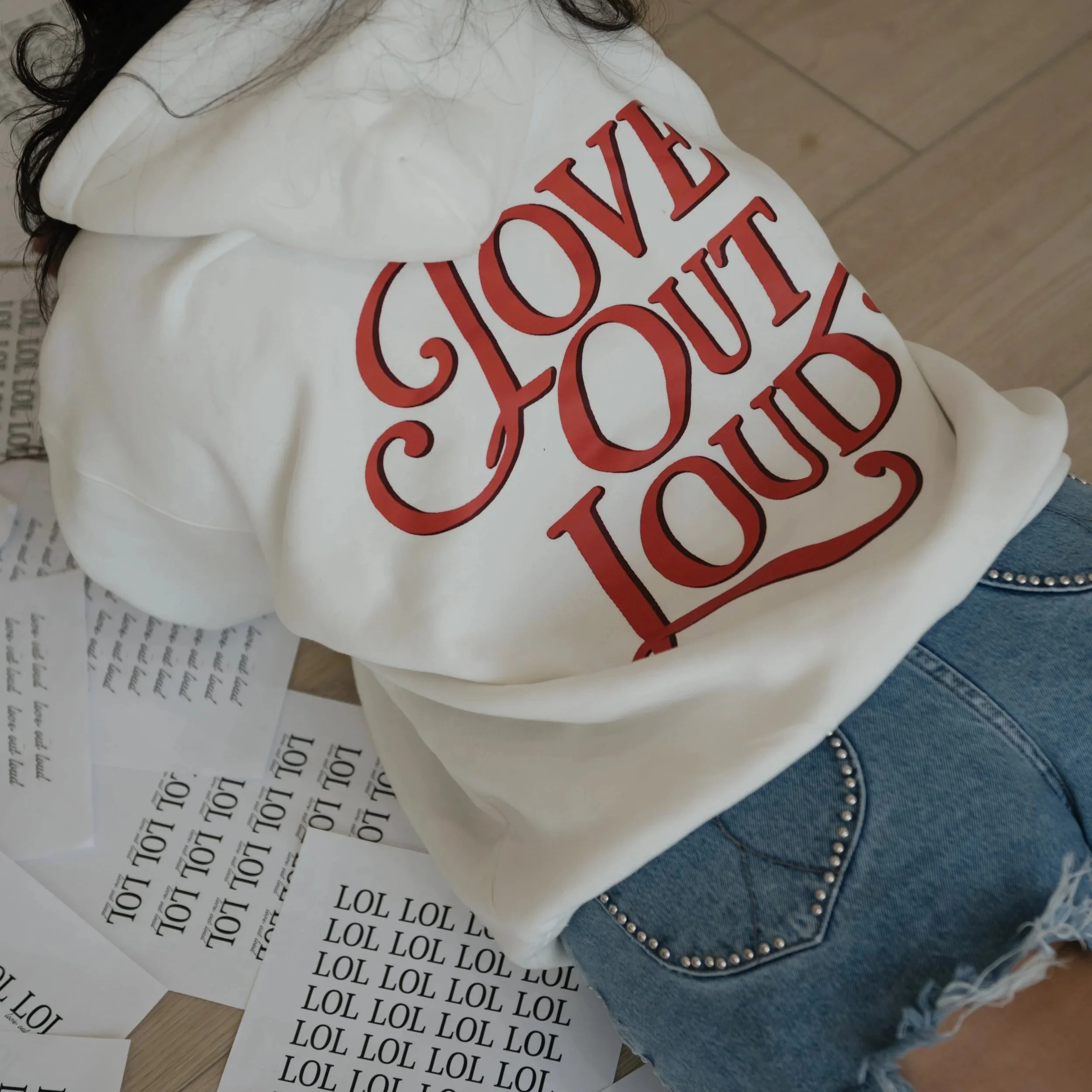 Piece of white clothing with large red text reading 'LOVE' on the chest area, denim jeans with silver embellishments, and scattered sheets of paper with repeated black text saying 'LOL' and other smaller text.