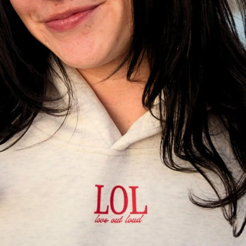 Close-up of a woman’s face smiling, showing part of her lips and chin, with dark hair, wearing a white shirt with red text that says 'LOL love out loud'.