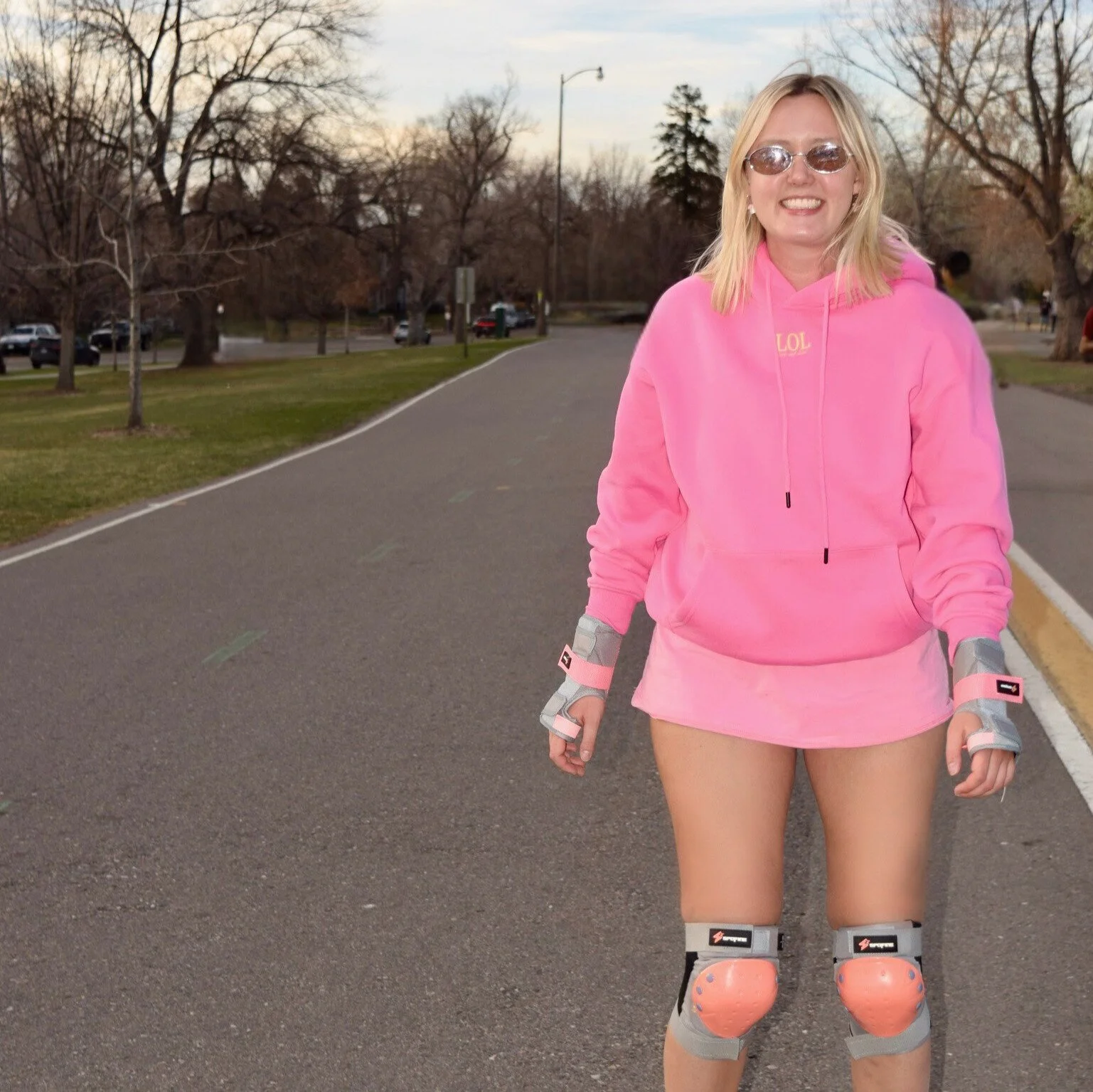 A young woman smiling outdoors in a pink hoodie and shorts, wearing protective gear on her elbows and knees, standing on a paved road with trees and parked cars in the background.