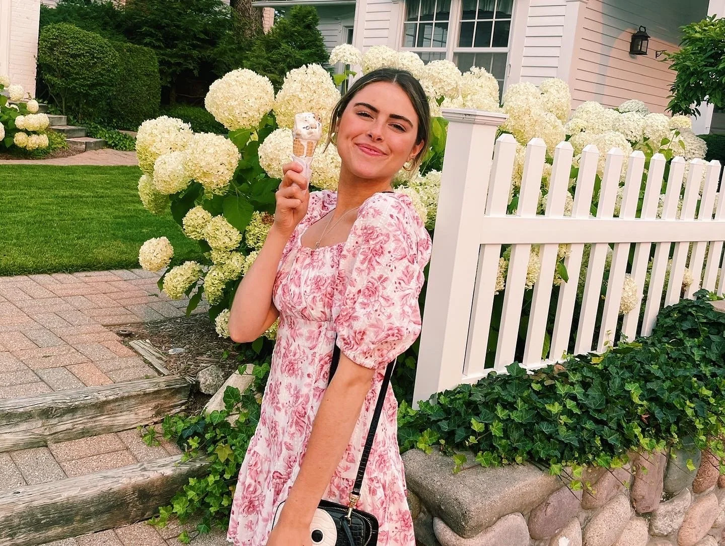 A young woman holding an ice cream cone in front of white hydrangea flowers and a white picket fence, smiling with closed eyes, wearing a pink floral dress, in a backyard with green grass and shrubs.