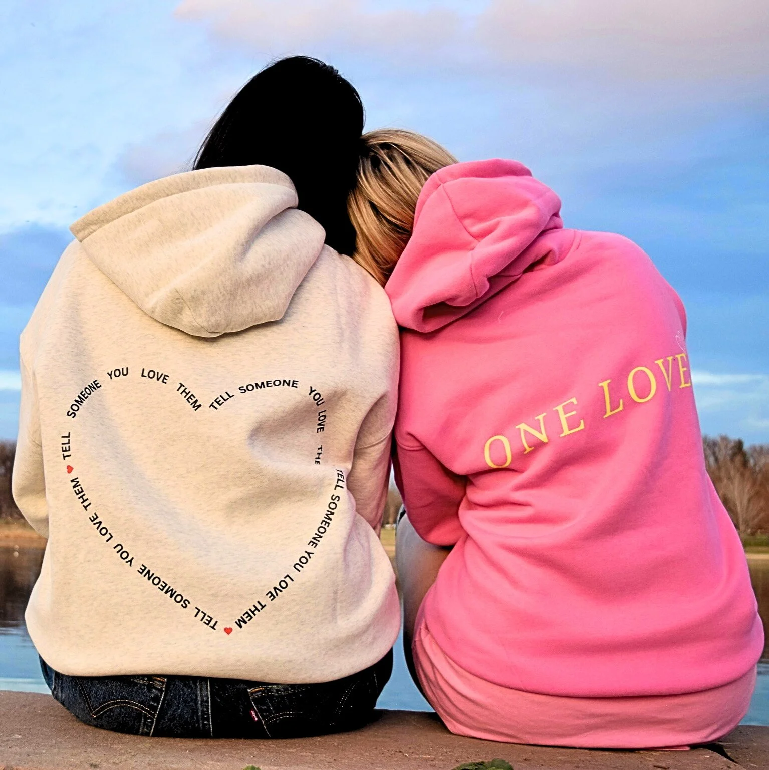 Two women sitting outdoors on a dock at sunset, wearing hoodies with love-themed messages, leaning on each other for support, near a lake with trees in the background.
