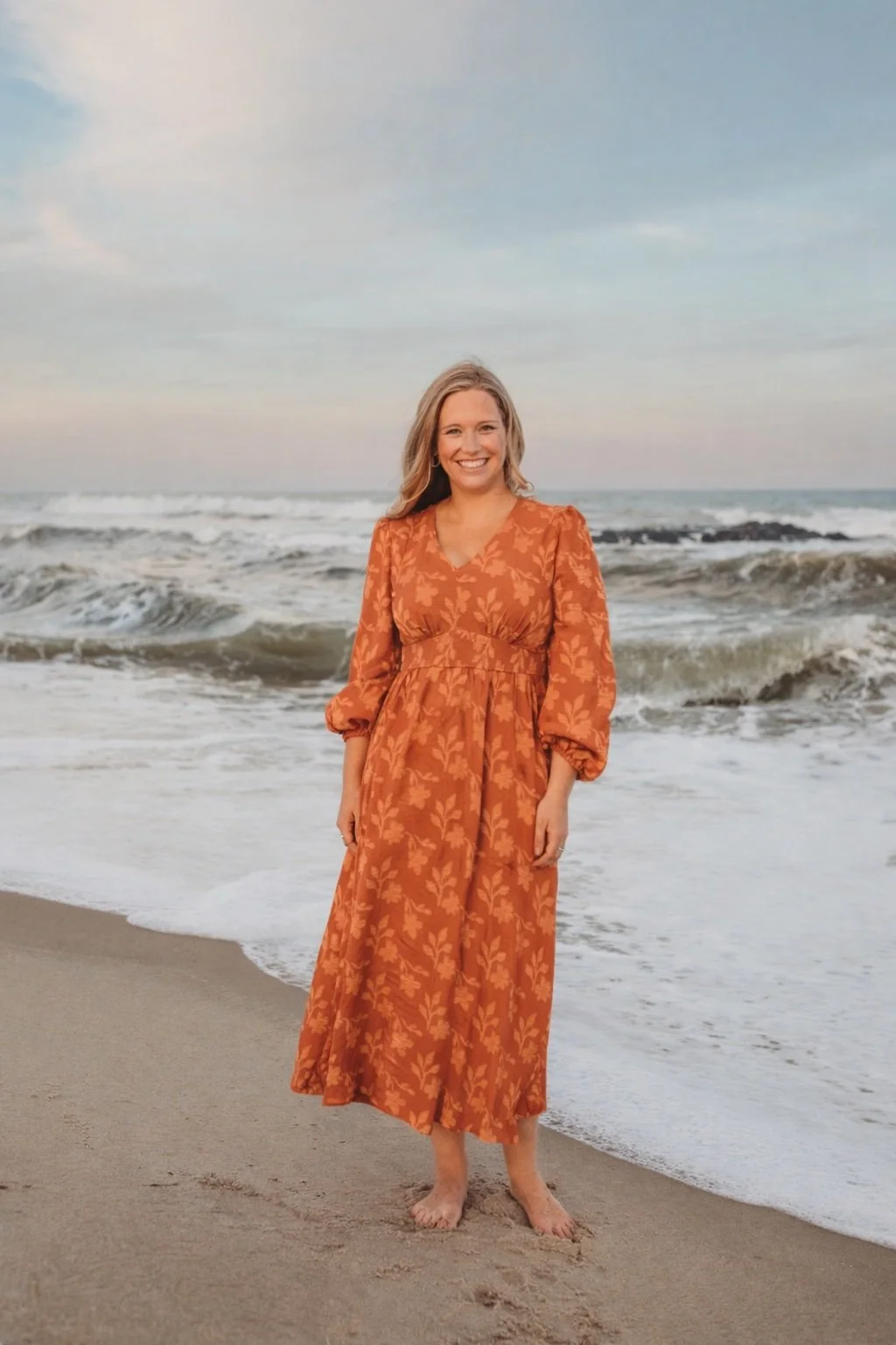 A woman standing on the beach with waves behind her. She is smiling, wearing an orange dress with a leaf pattern, and is barefoot.