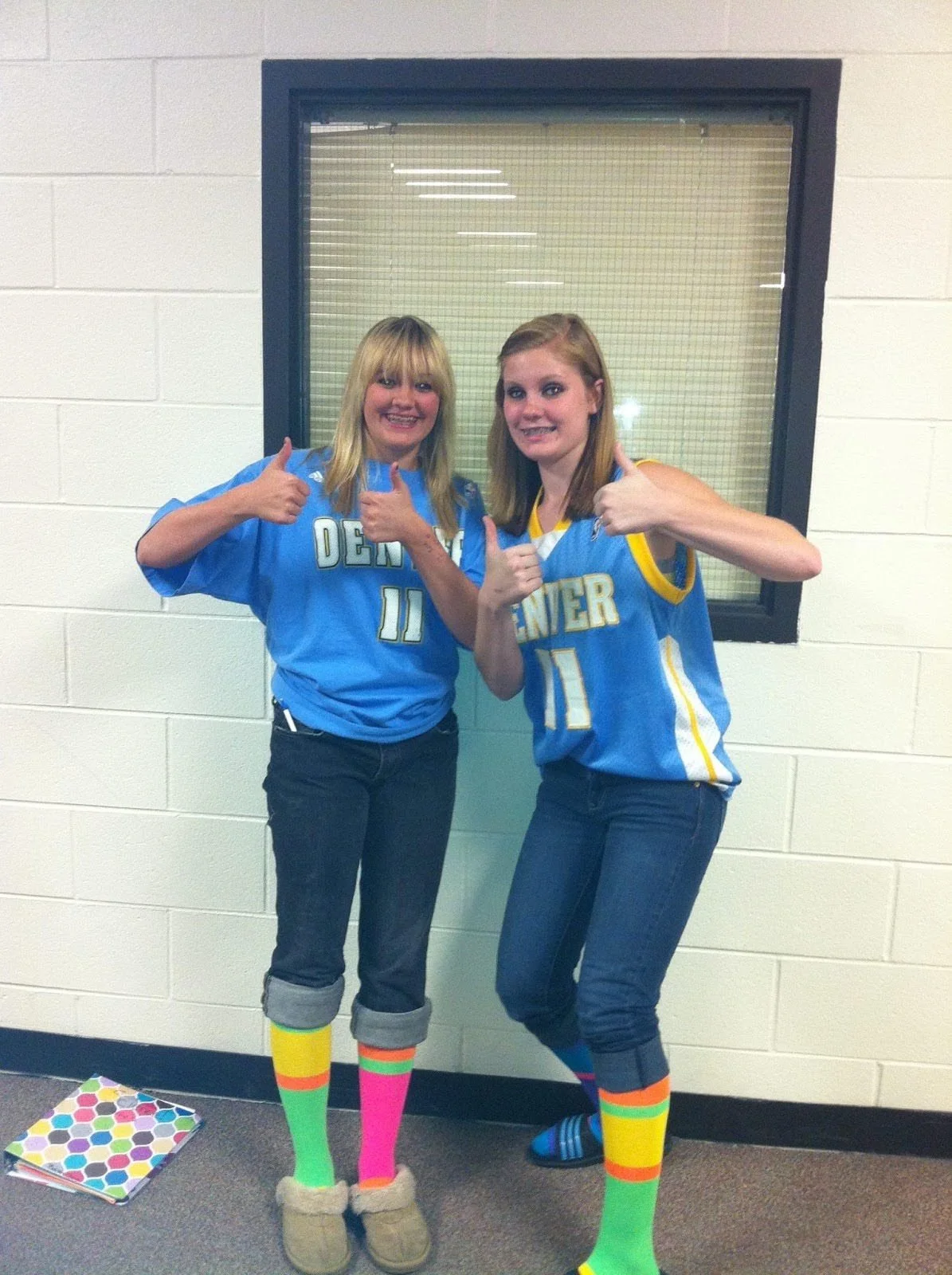 Two young women in matching blue Denver Nuggets jerseys giving thumbs up, wearing colorful striped socks, standing against a beige brick wall.