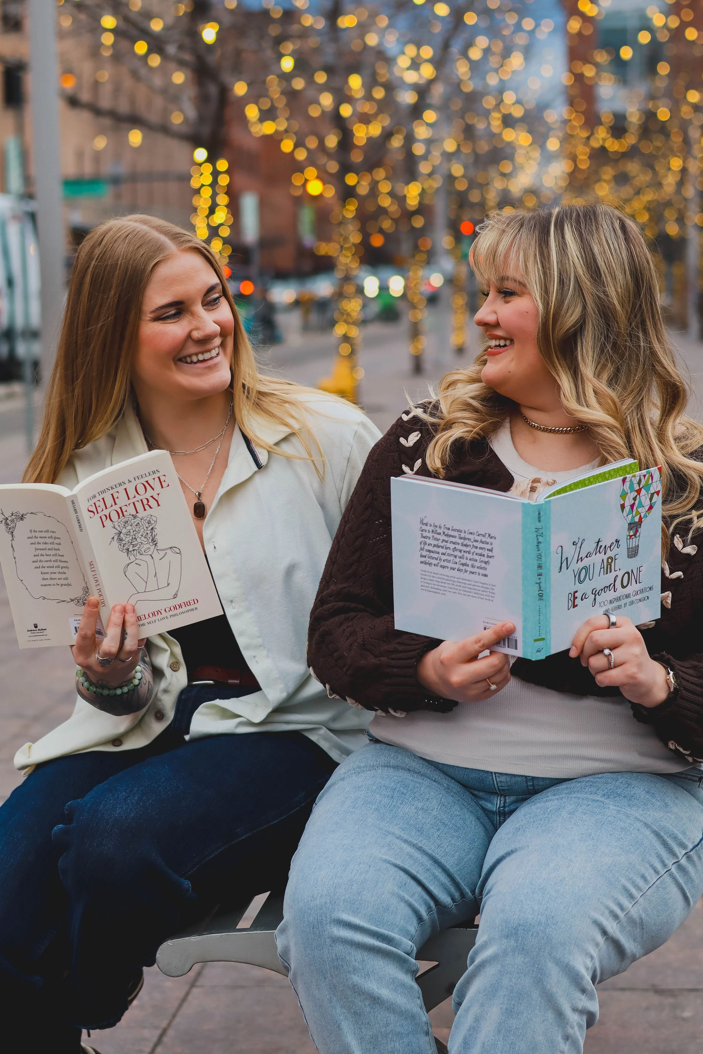 Two women sitting together outdoors laughing, each holding open books — "Self Love Poetry" and "Whatever You Are, Be a Good One."