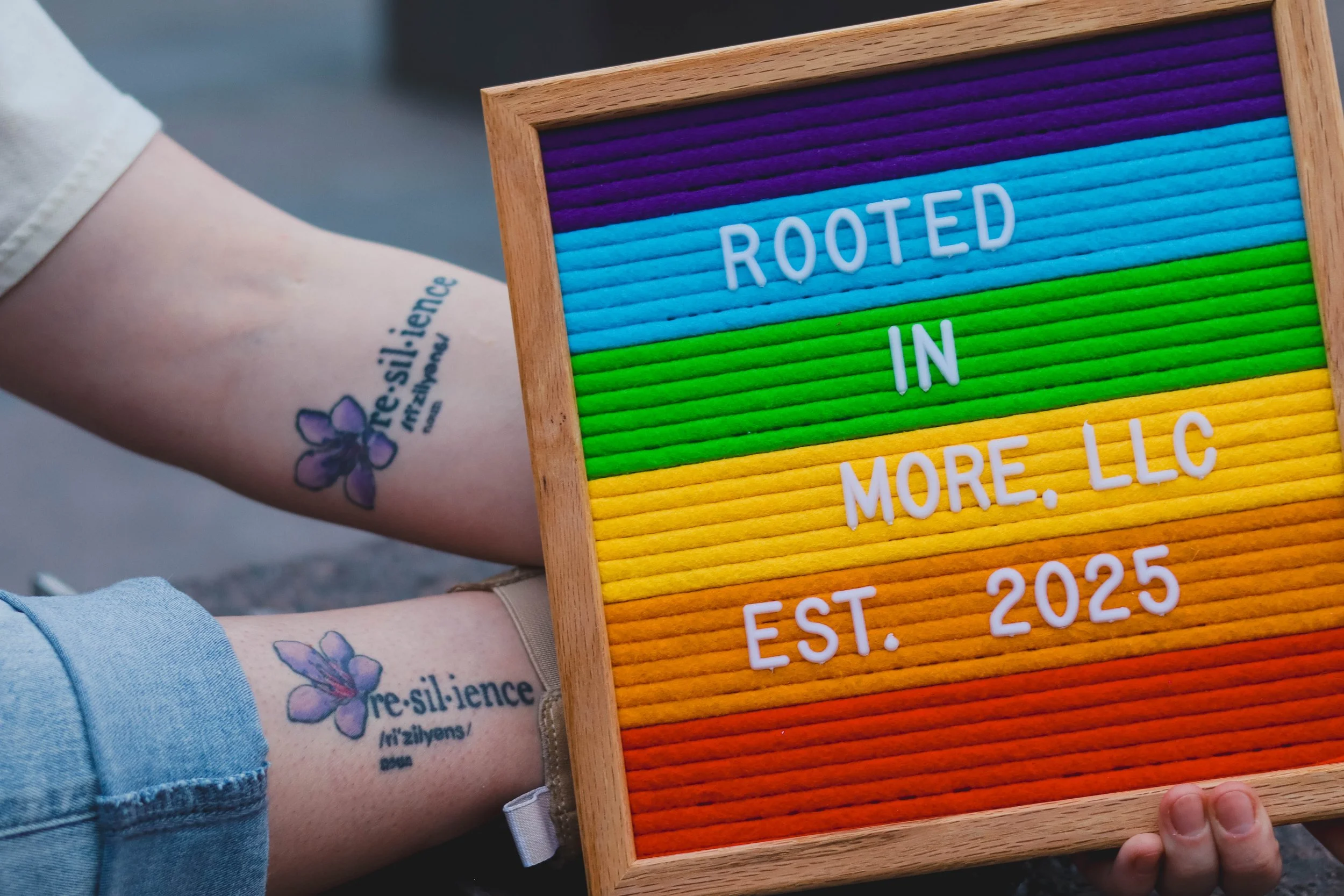 Person holding a colorful letterboard with the words 'Rooted in More, LLC' and 'Est. 2025' showing, and a visible tattoo of a Purple Saxifrage on their arm.