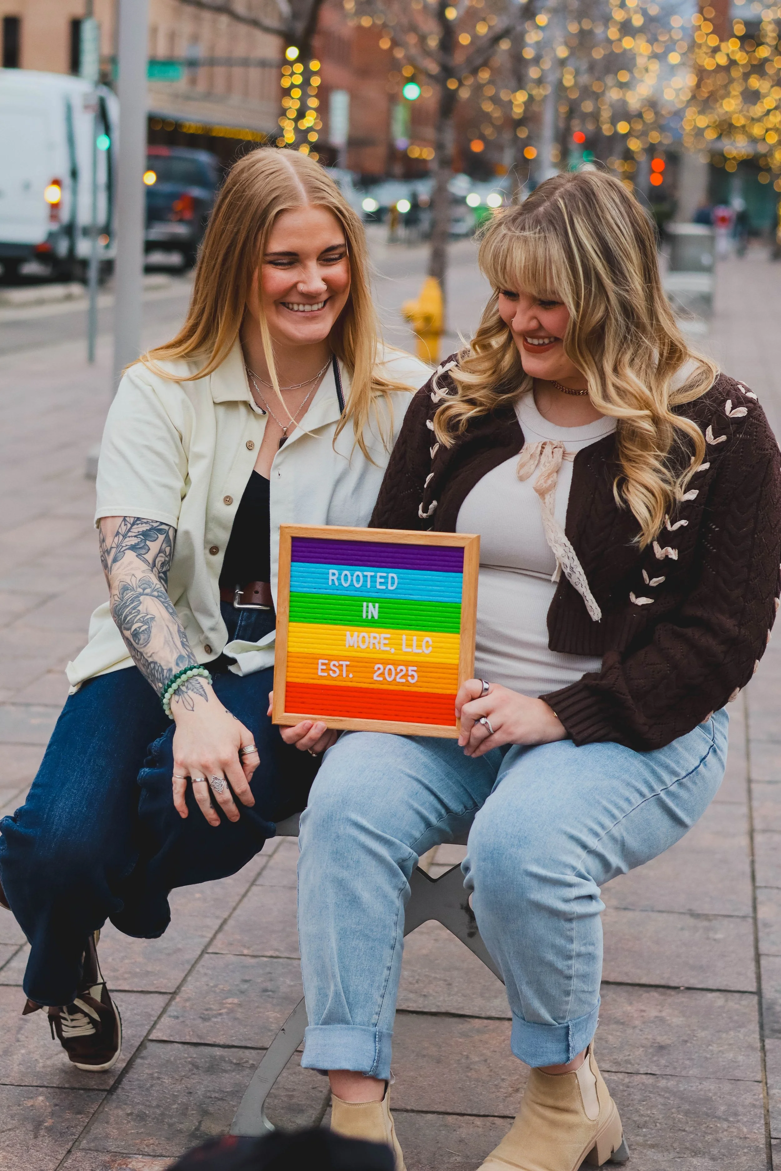 Two women sitting on a bench outdoors at dusk, smiling and looking at each other, holding a colorful sign that says 'Rooted in More, LLC Est. 2025.' The background features blurred city lights and trees with lights.