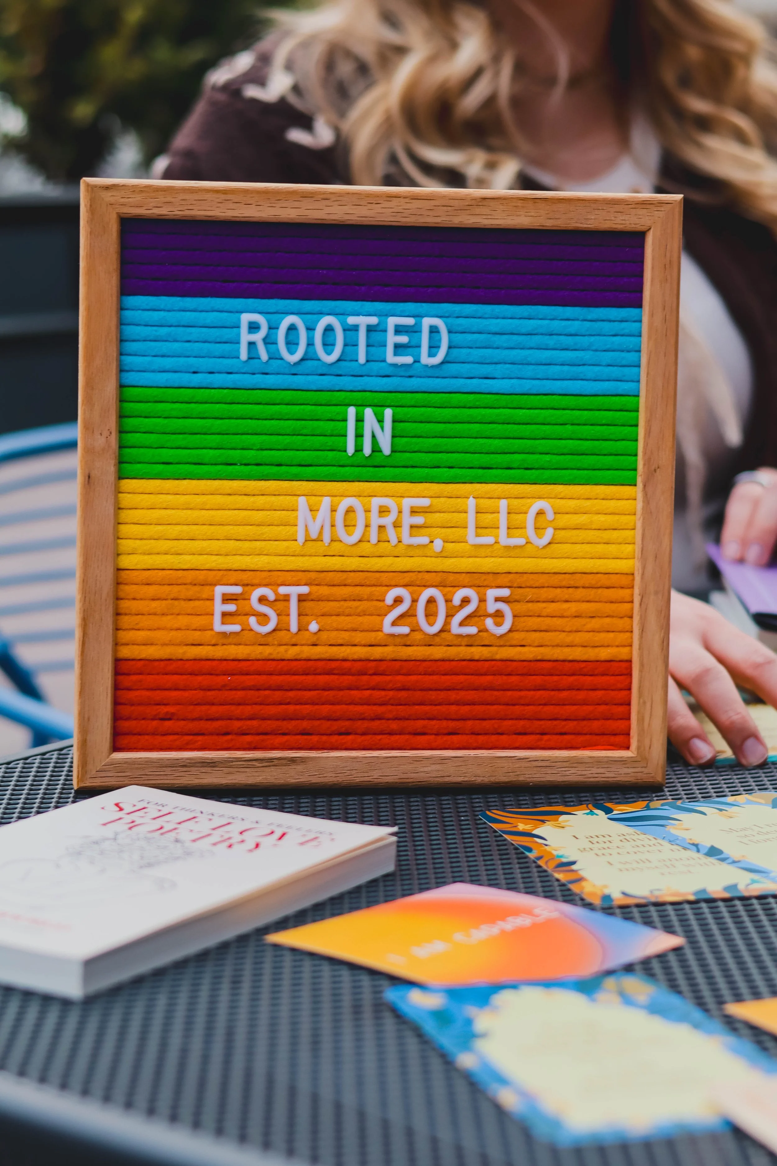 Close-up of a rainbow letter board reading "Rooted in More, LLC Est. 2025" resting on a table alongside books.