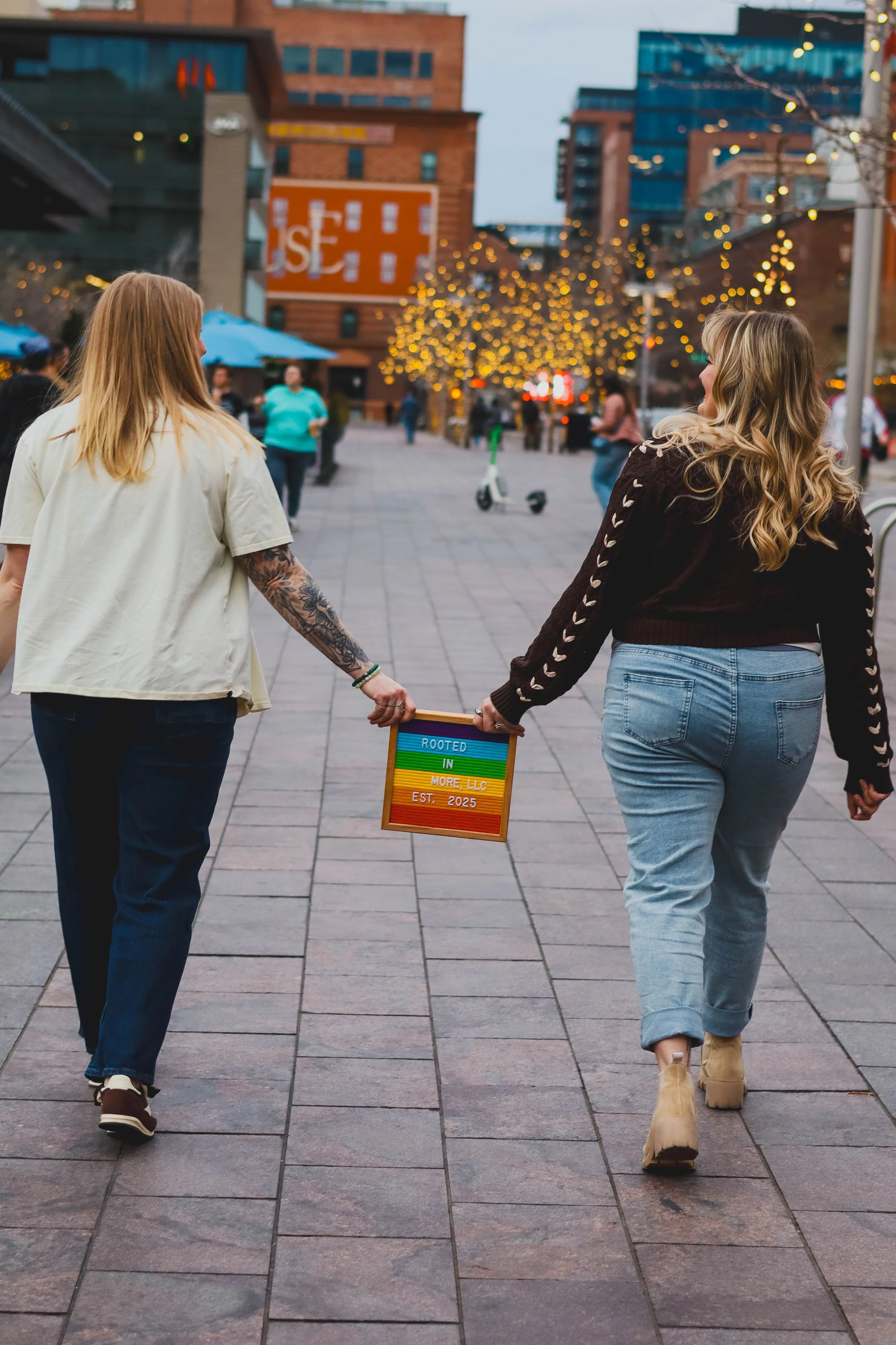 Two women walking hand-in-hand through an urban area with buildings and illuminated trees in the background. One woman has long blonde hair and is wearing a cream-colored shirt and dark jeans, while the other has wavy blonde hair and is dressed in a 