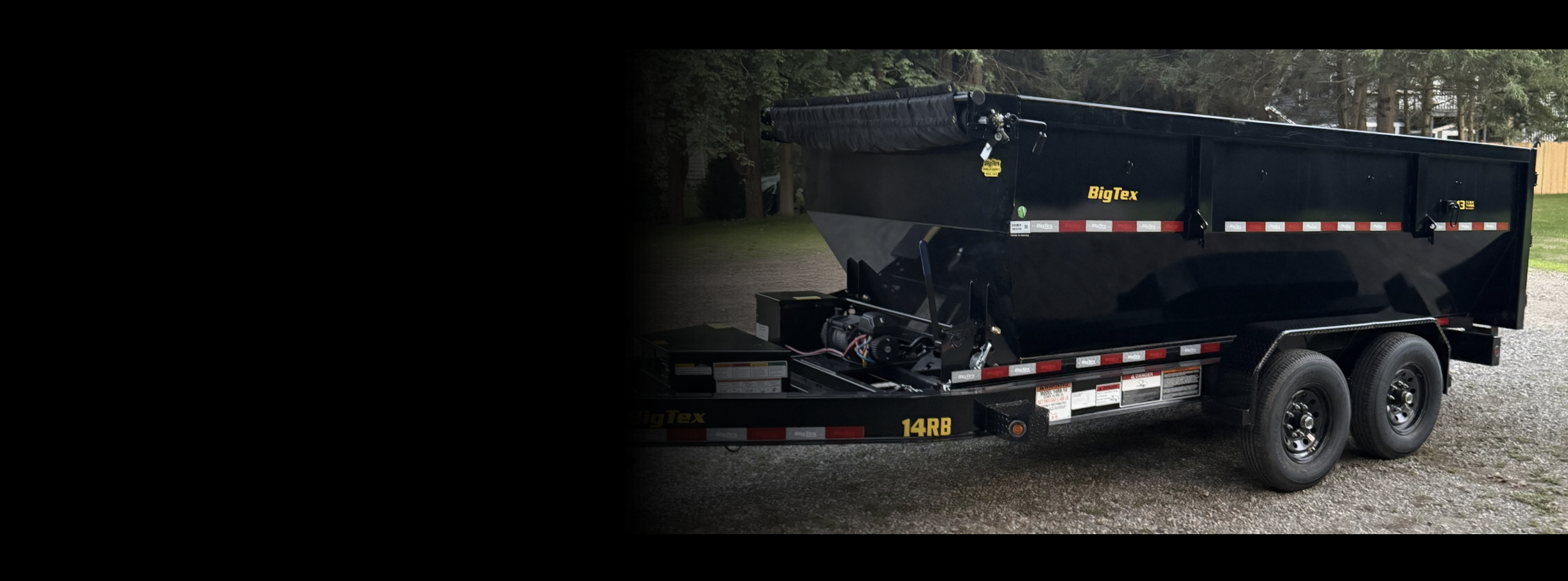 A large black Big Tex dump trailer with dual axles and tires parked on a gravel lot, with trees in the background.