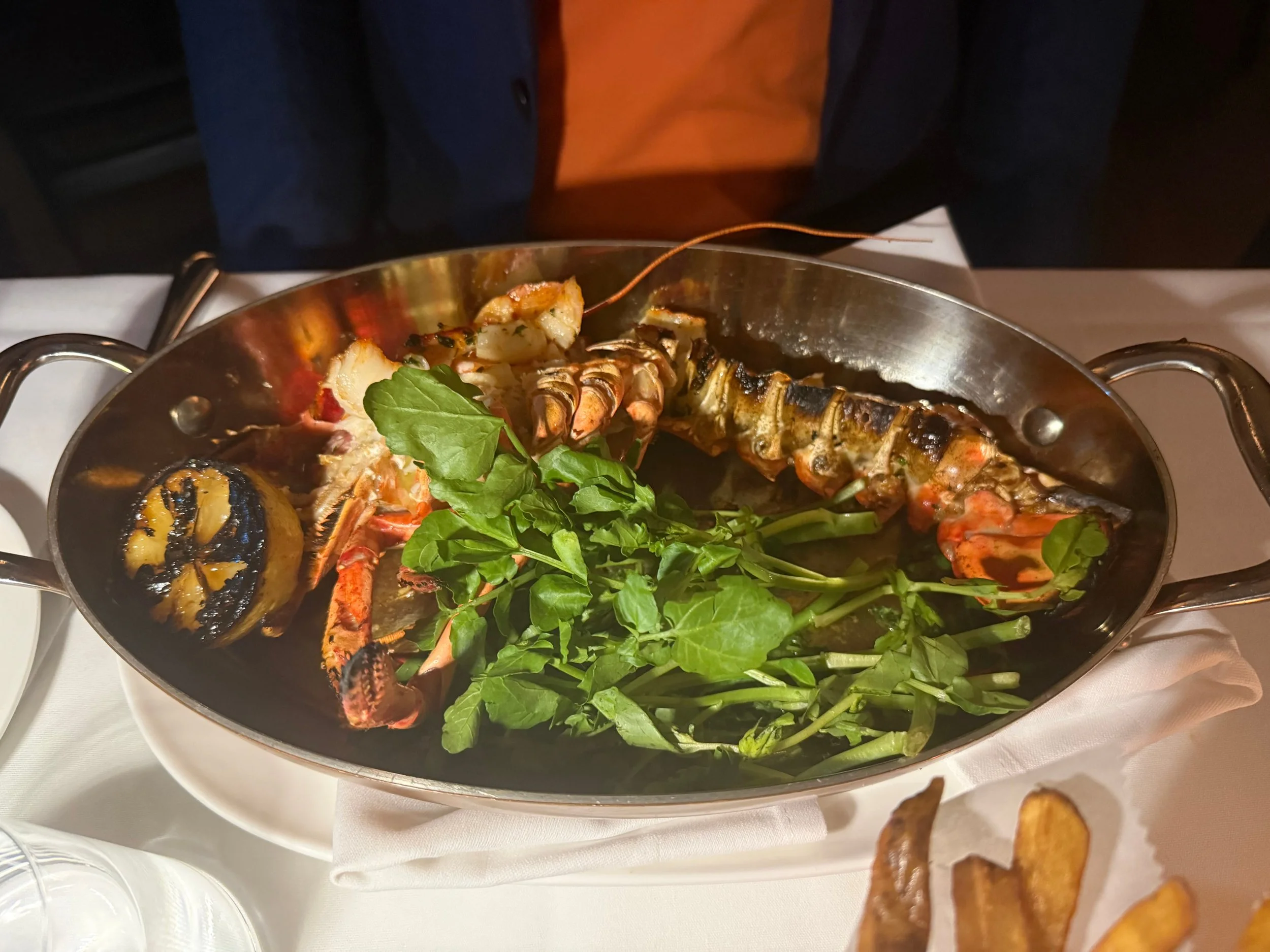 A surf and turf dish featuring grilled lobster tail and a large lobster claw, accompanied by fresh green herbs and served in a large metallic pan.