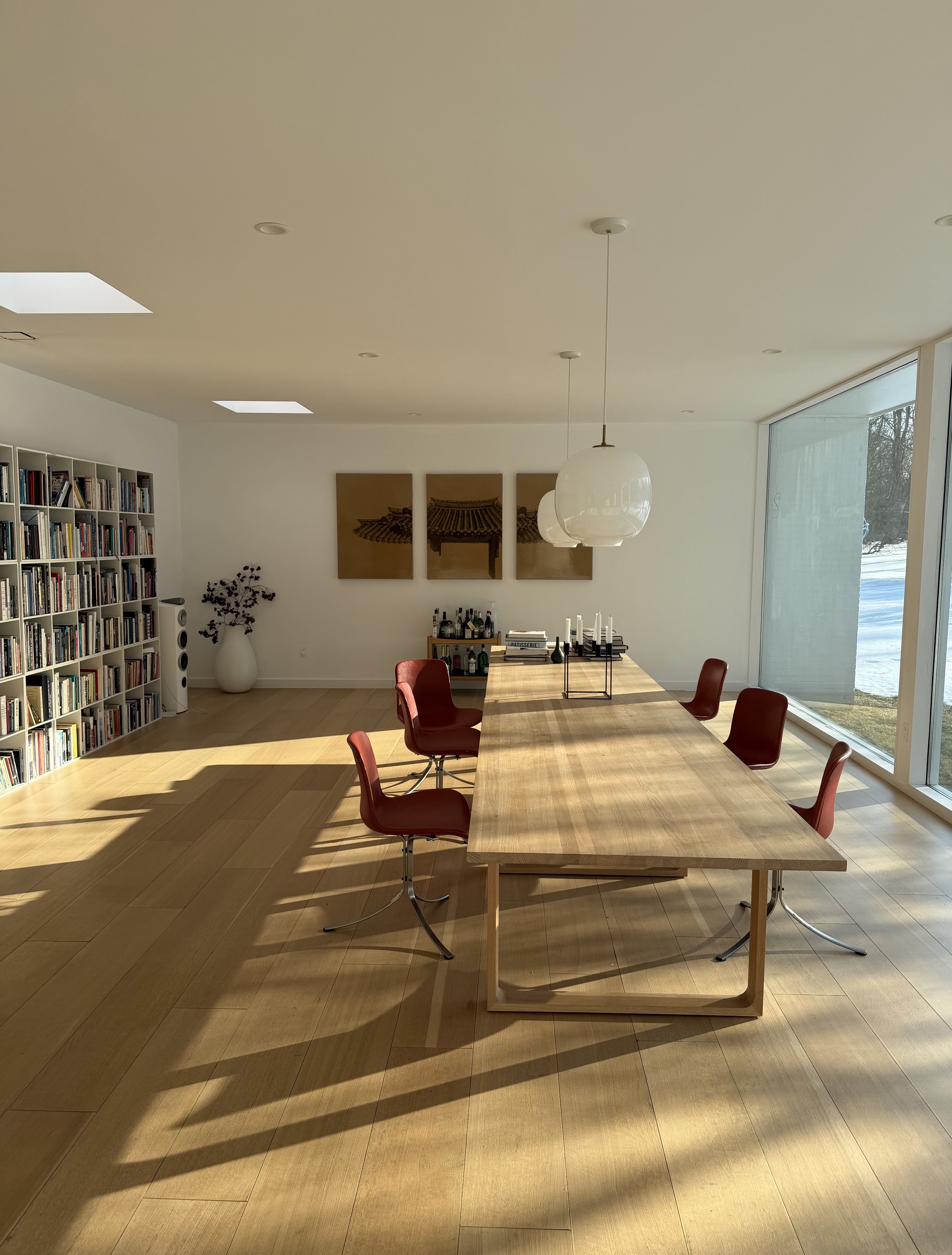 Modern dining room with a wooden table and six red chairs, large window with outdoor view, bookshelf on the left, art on the back wall, and ceiling lights.
