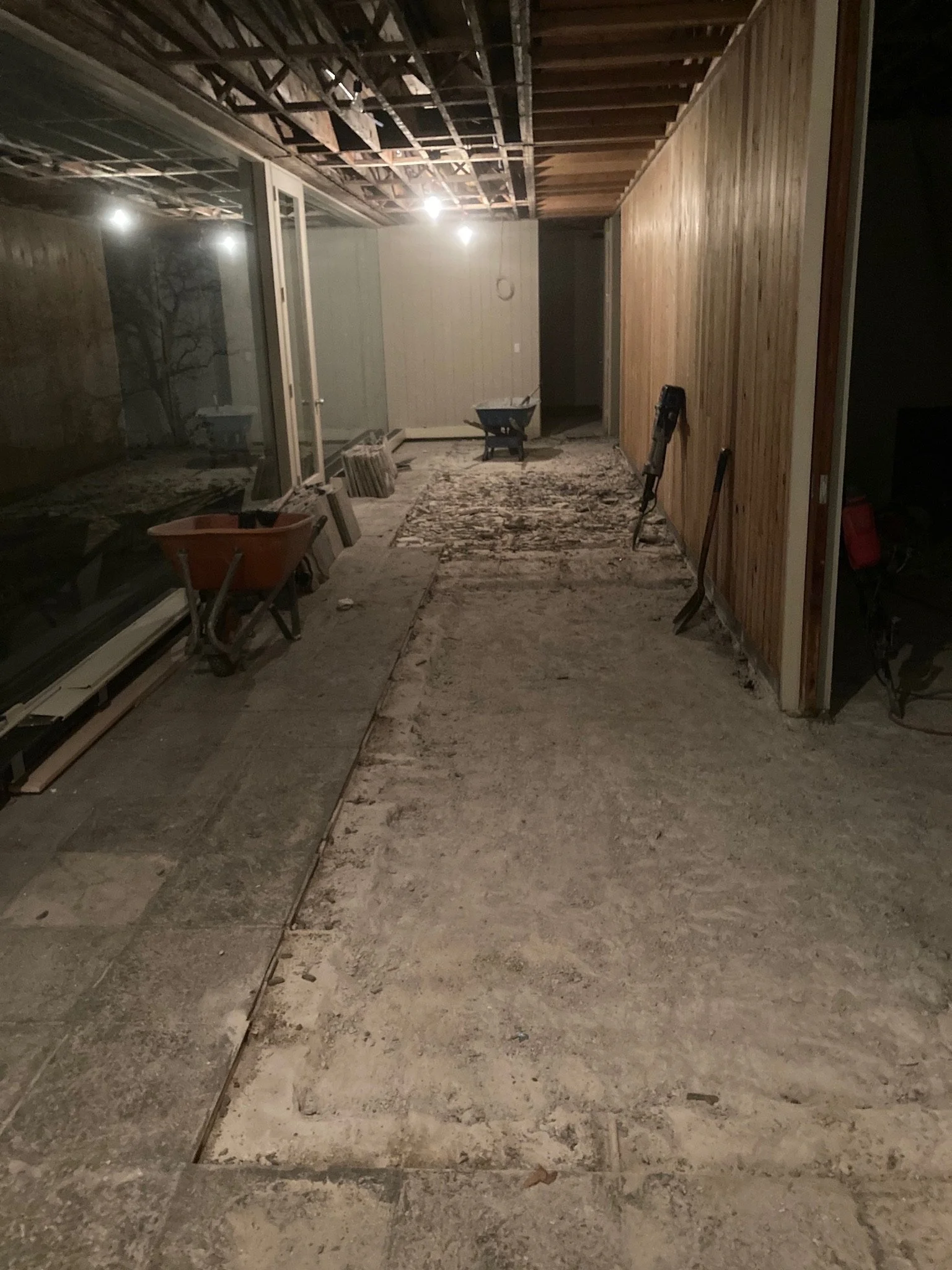 A room under construction with exposed ceiling beams, unfinished flooring, and construction tools such as a wheelbarrow, shovel, and saw. There is a large glass window on the left side, and a partially paneled wooden wall on the right.