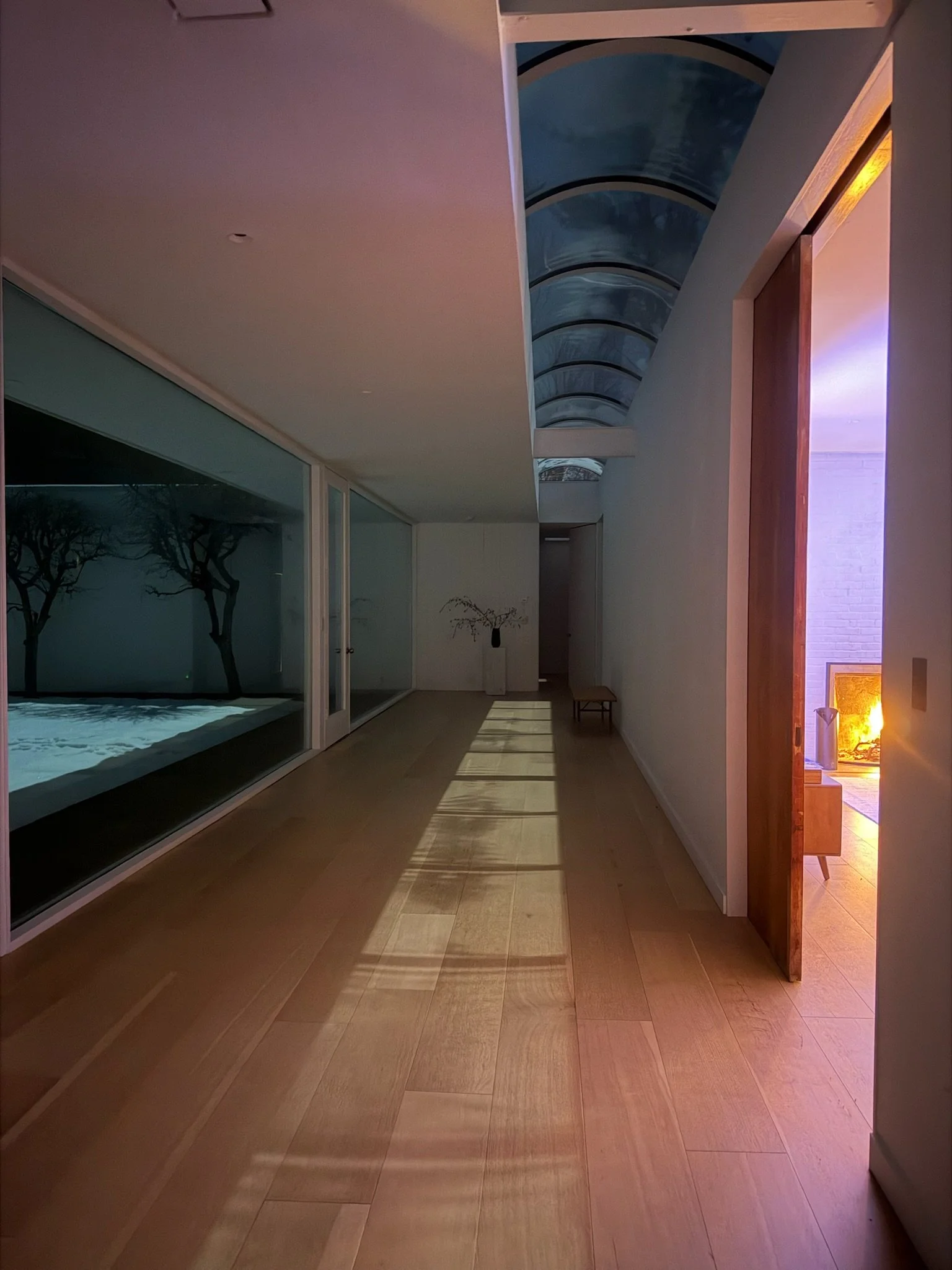 Interior view of a modern hallway with wooden floors, large glass window on the left, a hallway with shadows cast on the floor, a doorway on the right leading to a lit room, and a curved glass ceiling allowing natural light.