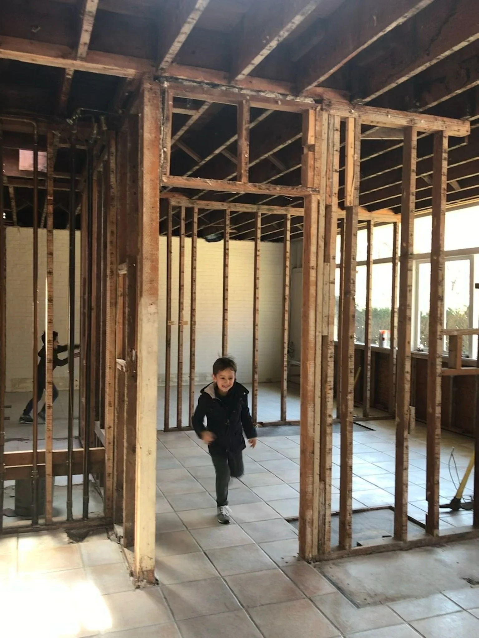 A young boy running through the frame of an unfinished room in a house under construction, with visible wooden framing and tiled flooring.
