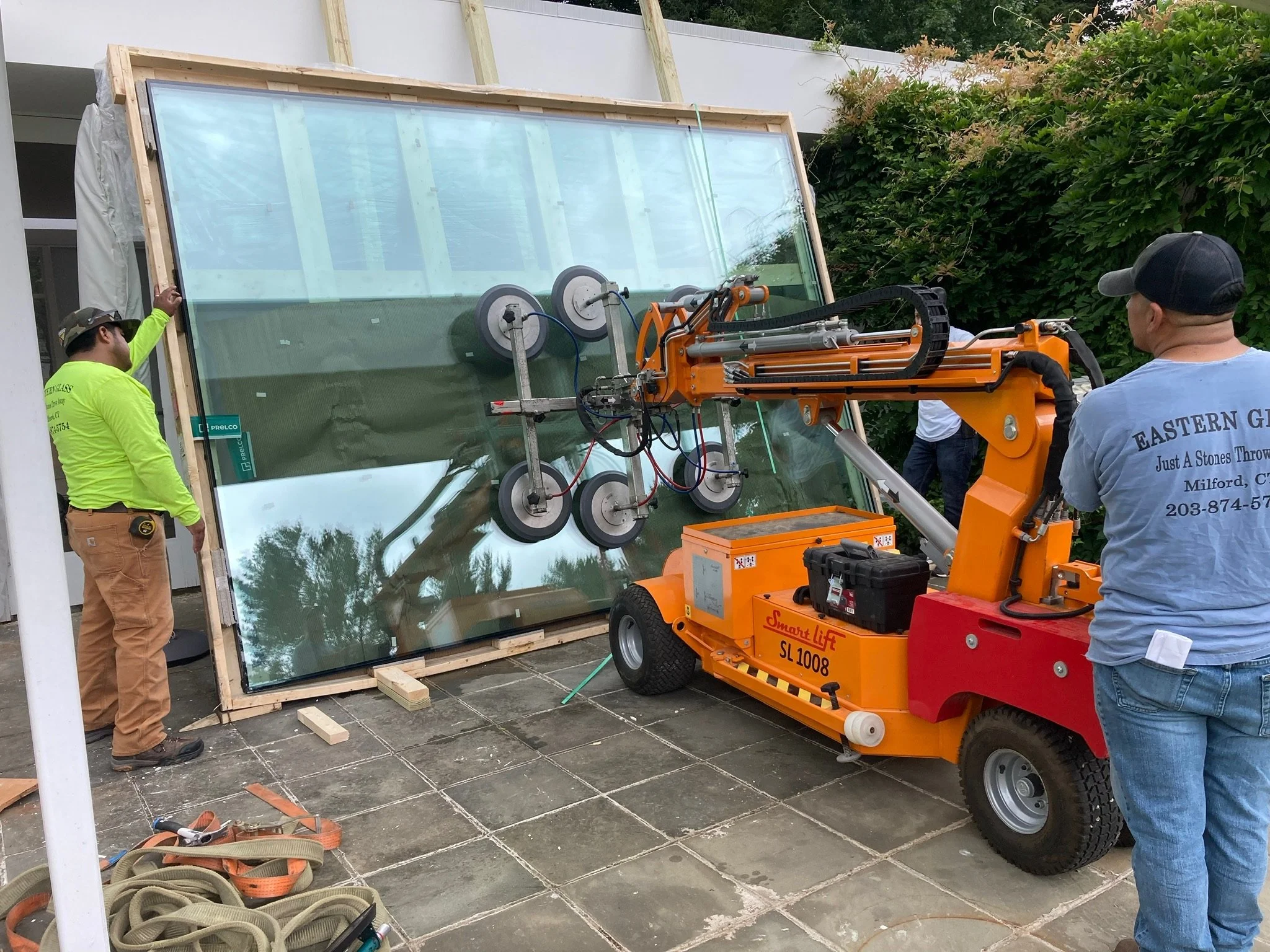 Workers installing a large glass window using a specialized robotic lifting machine on an outdoor patio.