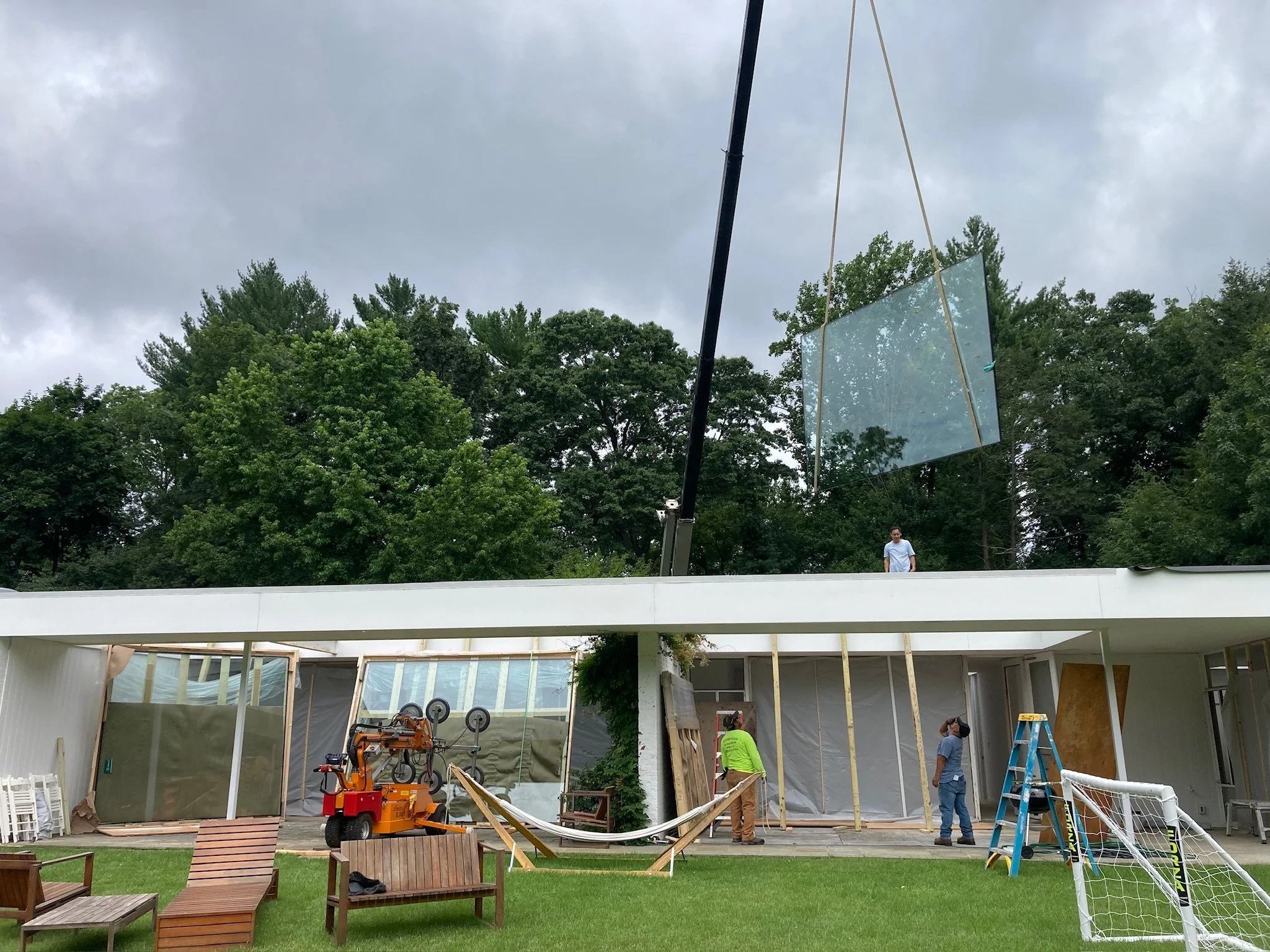 Construction workers building a modern house with large glass windows and an outdoor area with benches and a small goal post. A person is standing on the roof, and there's a crane lifting a large glass panel.
