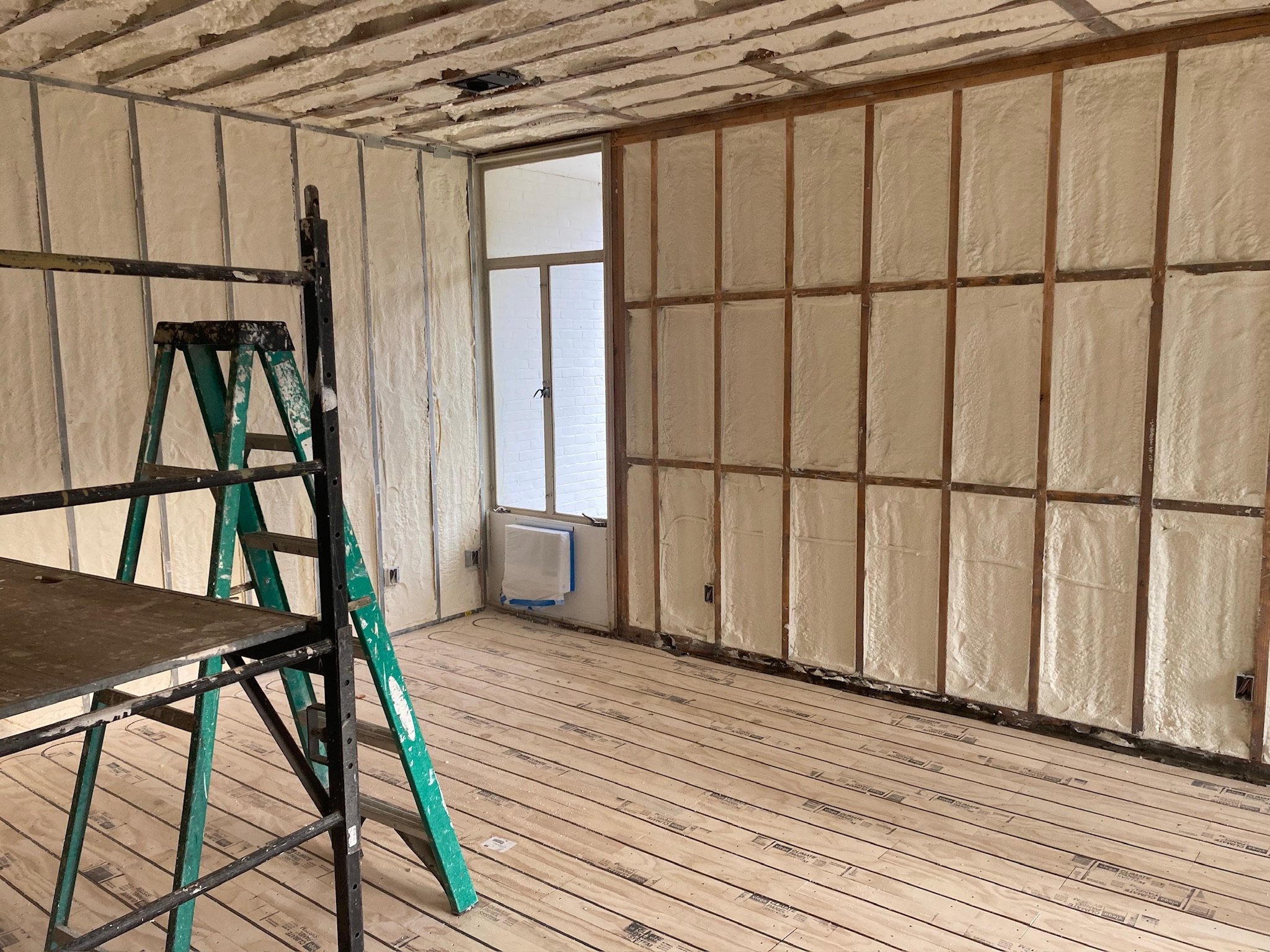 Interior of a room under renovation with insulation spray foam on the walls. There is a window, a green ladder, and construction materials on the floor.