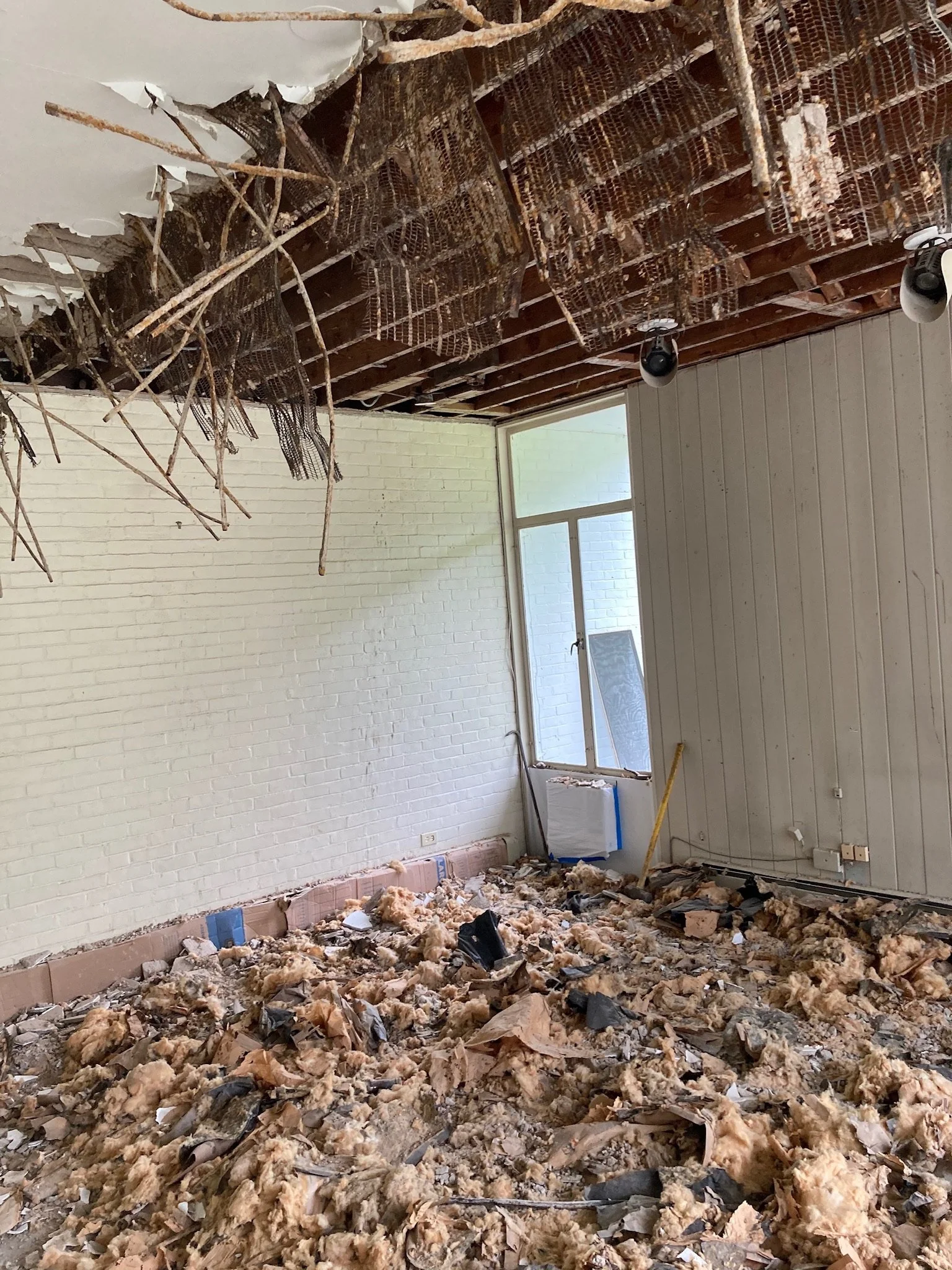 Room with ceiling damaged and insulation. Floor covered with debris, broken materials, and insulation. Window on the back wall.