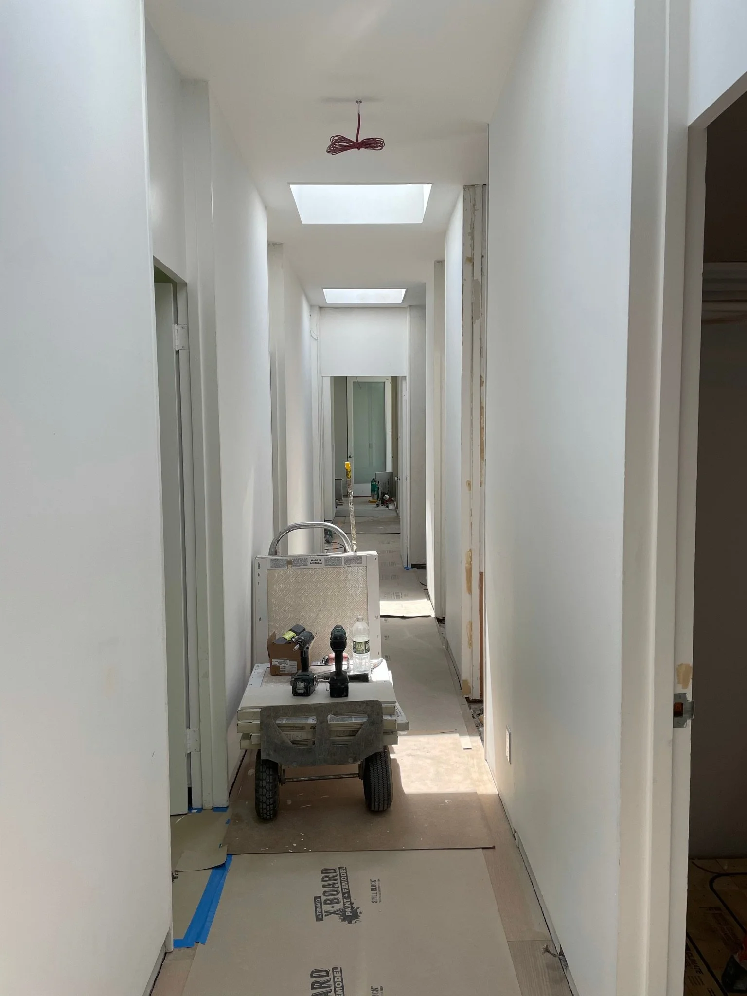 Construction hallway with white walls and ceiling, unfinished doorways, a cart with tools, and skylights on the ceiling