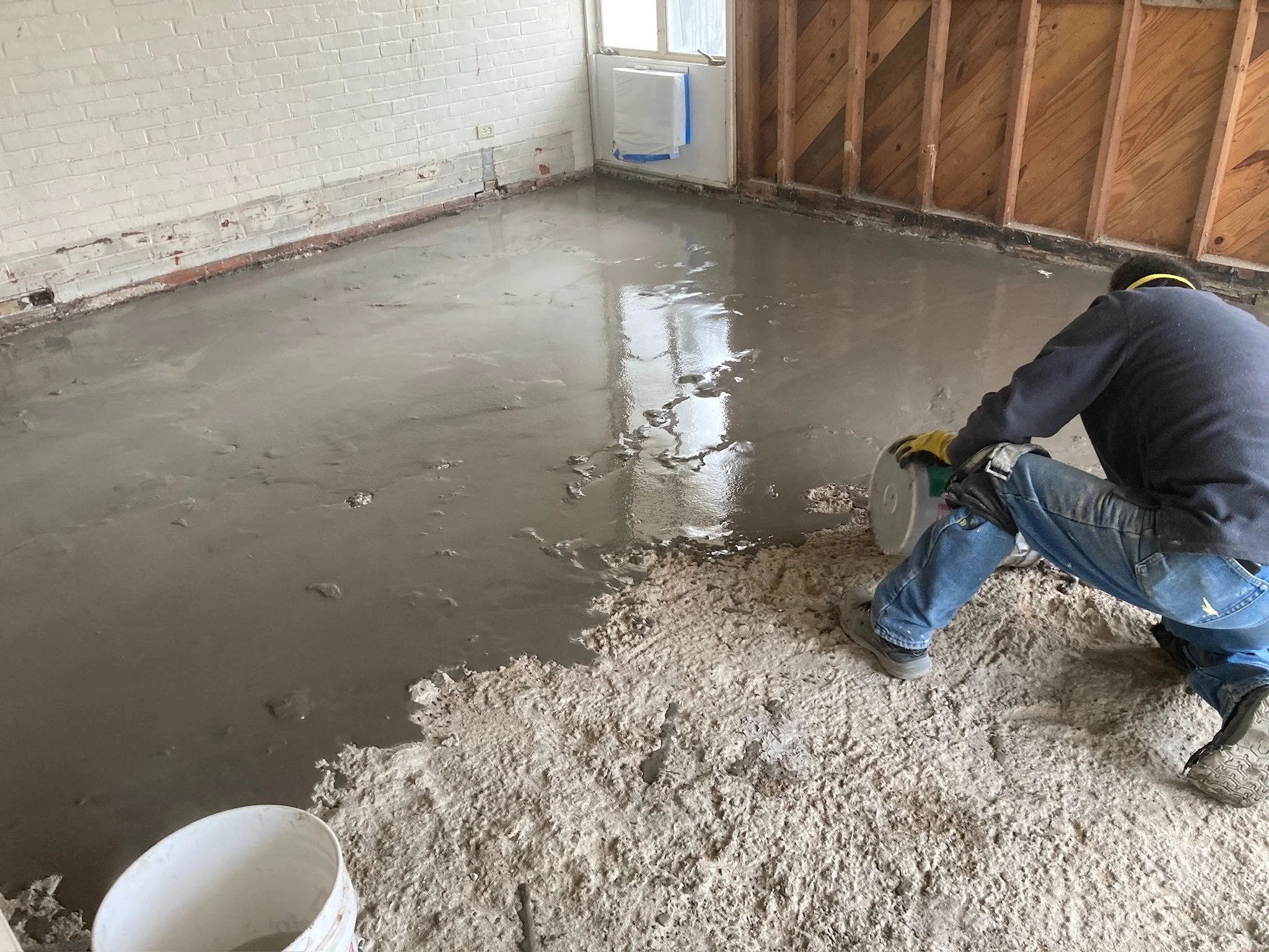 A worker is pouring wet concrete onto a floor inside a room with exposed wood and brick walls, and a window.