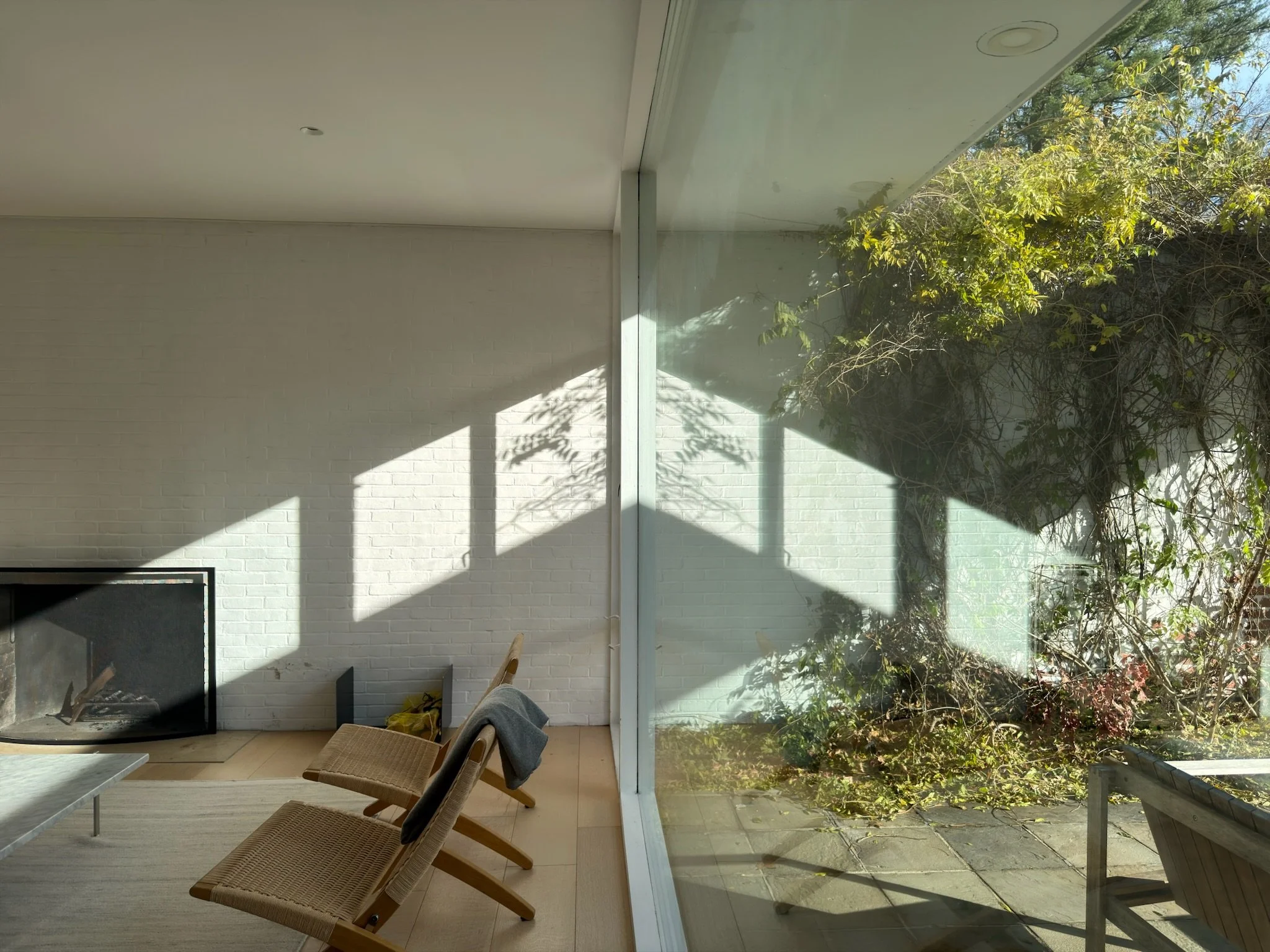 Living room with a white brick fireplace, two woven chairs with gray blankets, a glass wall with a view of outdoor greenery and patio, sunlight and shadows cast inside.