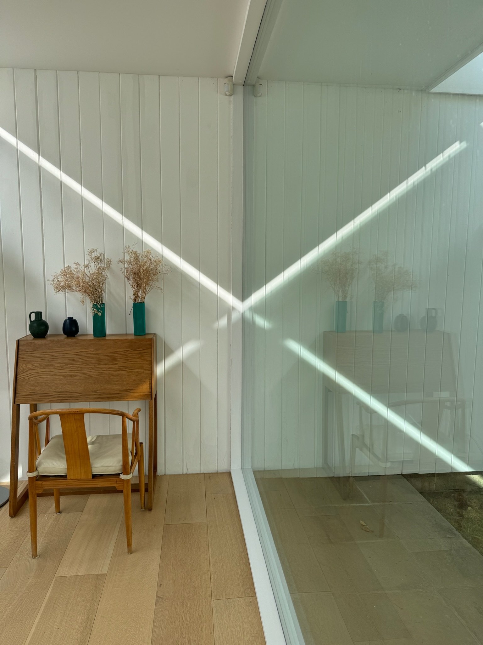 corner of room with wooden chair, small table, and three vases with dried flowers, next to glass wall with reflections and diagonal sunlight.