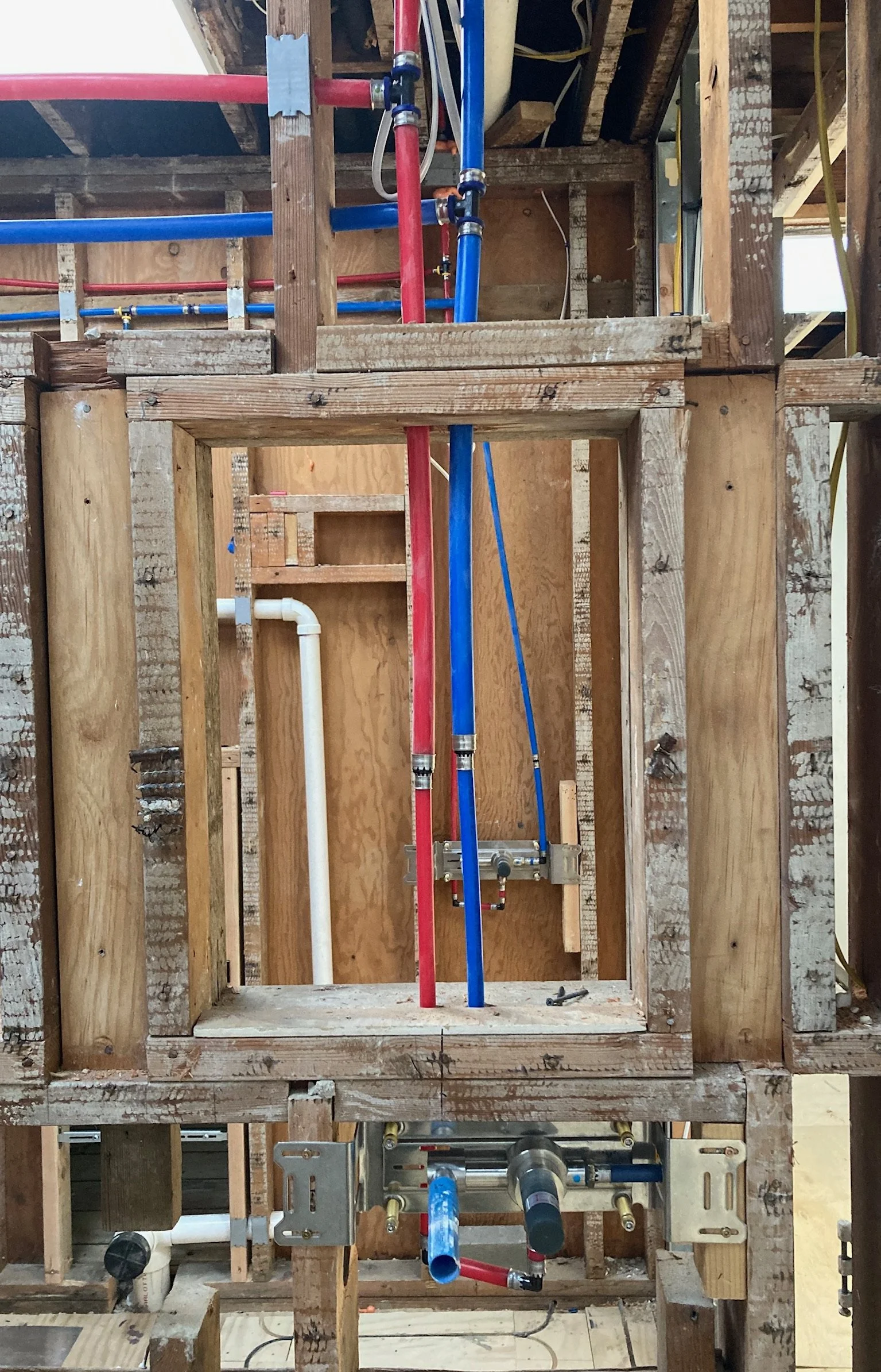 Construction framing with exposed wooden studs and plumbing pipes, including red, blue, and white pipes, in a building under construction.