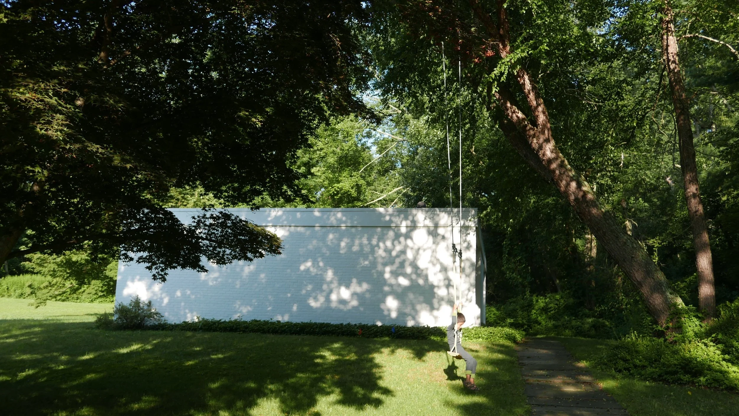 Child swinging on a swing set in a grassy yard shaded by large trees, with a white building in the background and dappled sunlight on the ground.