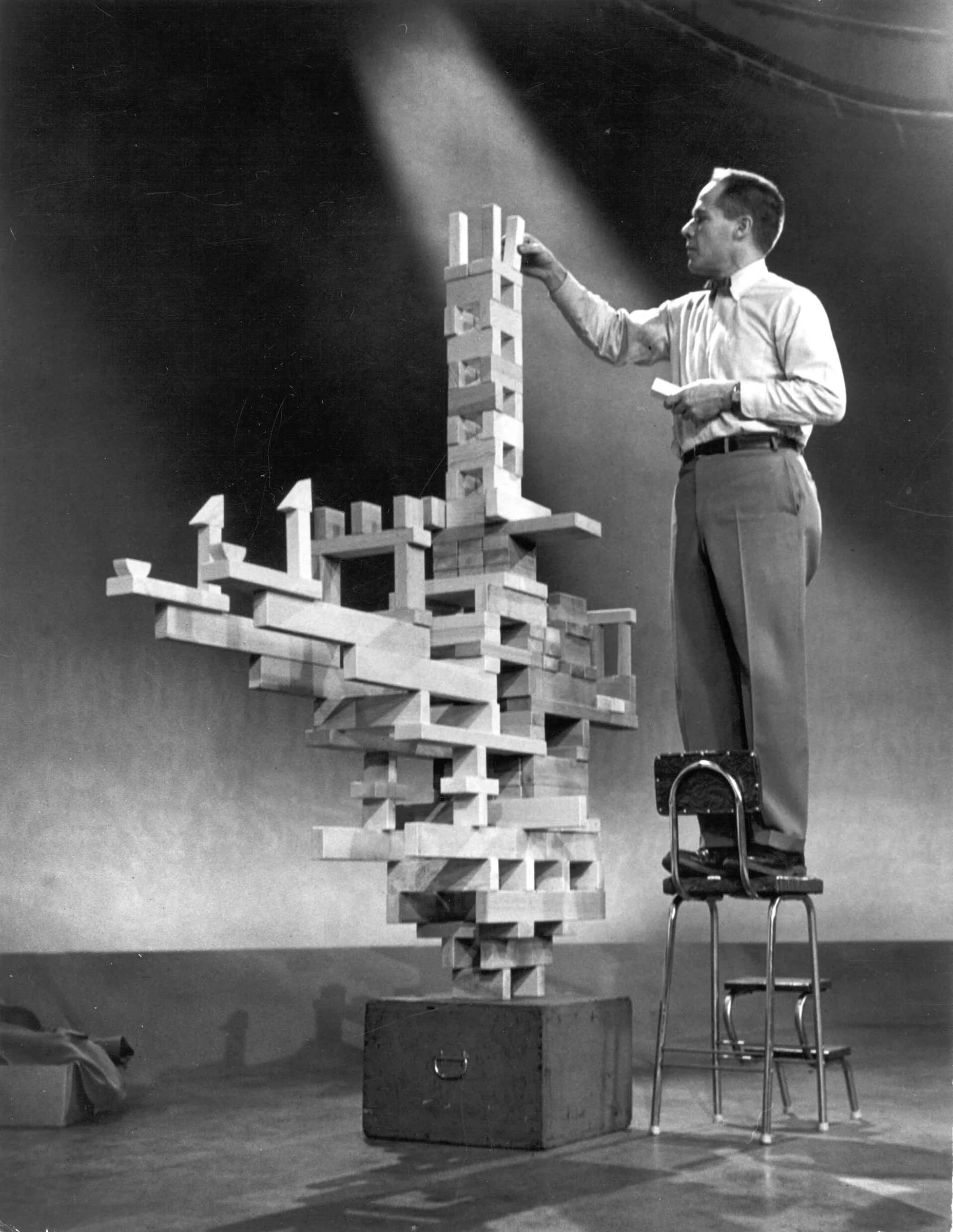 A man standing on a chair constructing a large, abstract wooden structure on a stage. The structure appears to be a modern art or architectural model with various interconnected blocks and platforms.