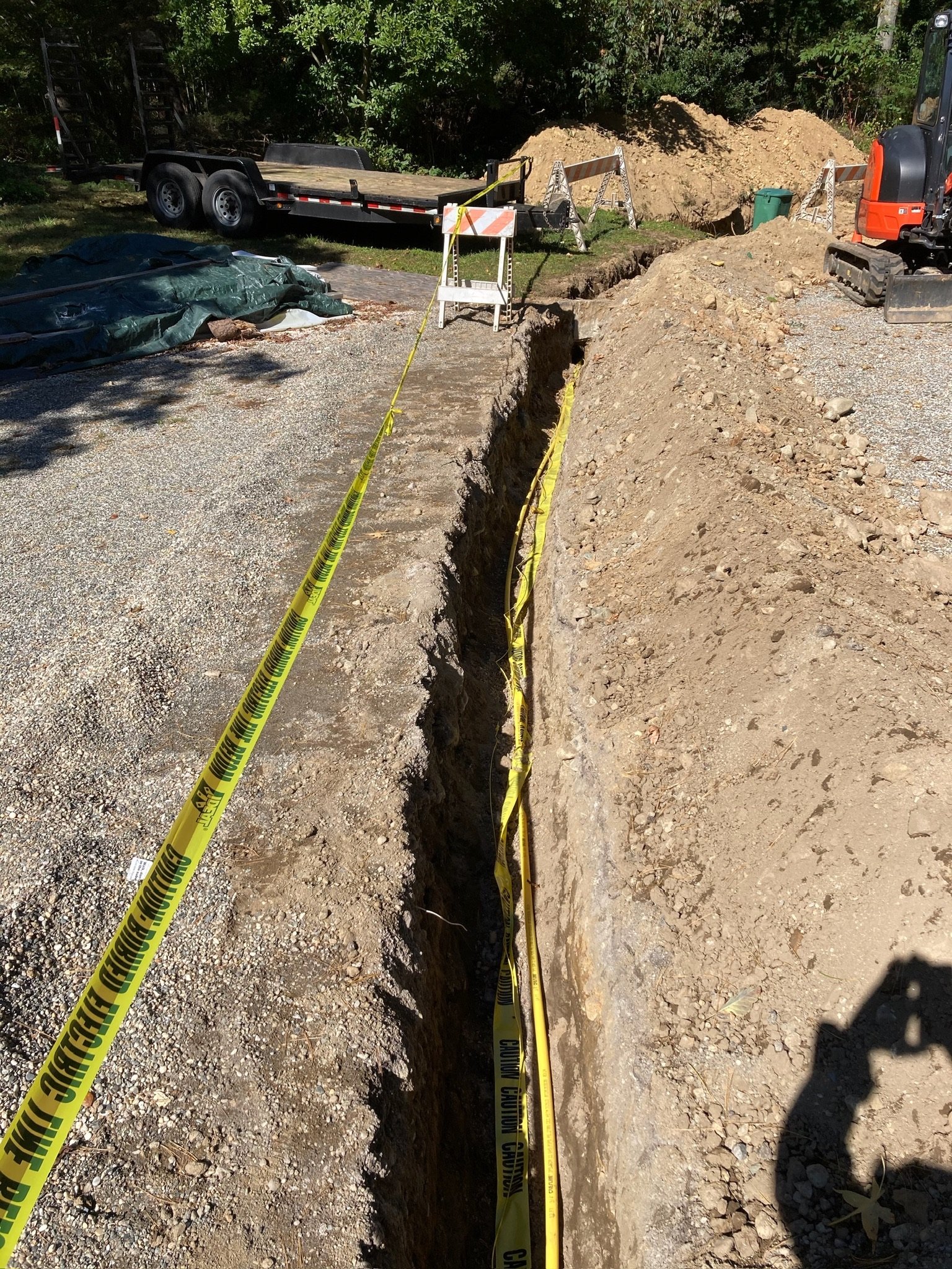 Construction site with a deep trench, caution tape, a small excavator, and construction barriers, surrounded by dirt and gravel.