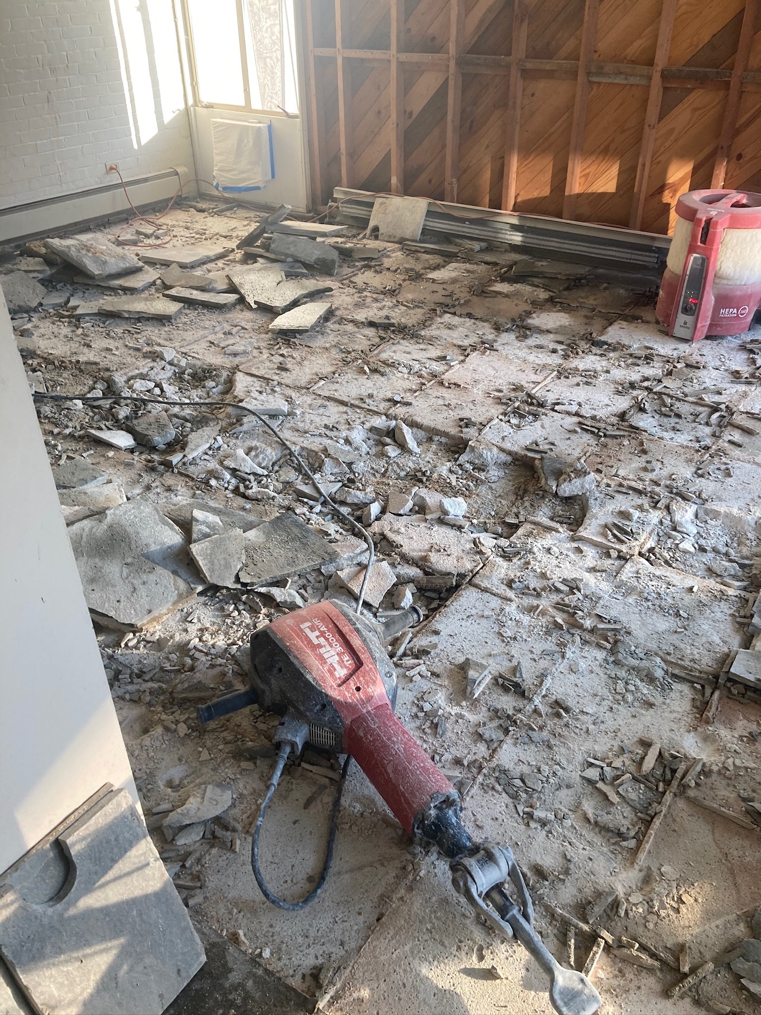 Room under renovation with removal of old flooring, debris scattered, and construction tools including a floor scraper on the dusty floor, partial wooden wall framing, a window with sunlight, and construction equipment in the corner.