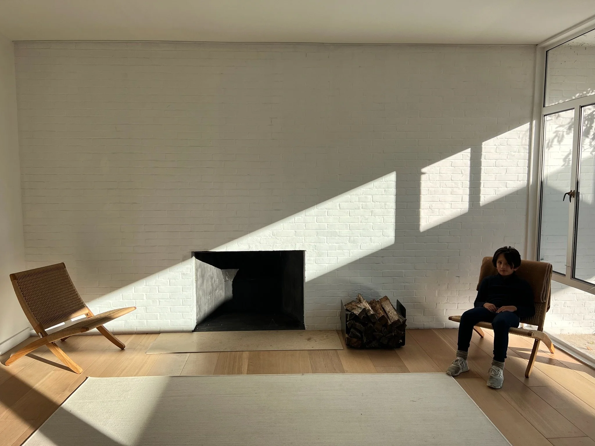 A minimalist living room with a white brick wall, a fireplace, a wood basket of logs, a person sitting in a brown chair, and sunlight streaming through large windows creating a pattern of light and shadow.