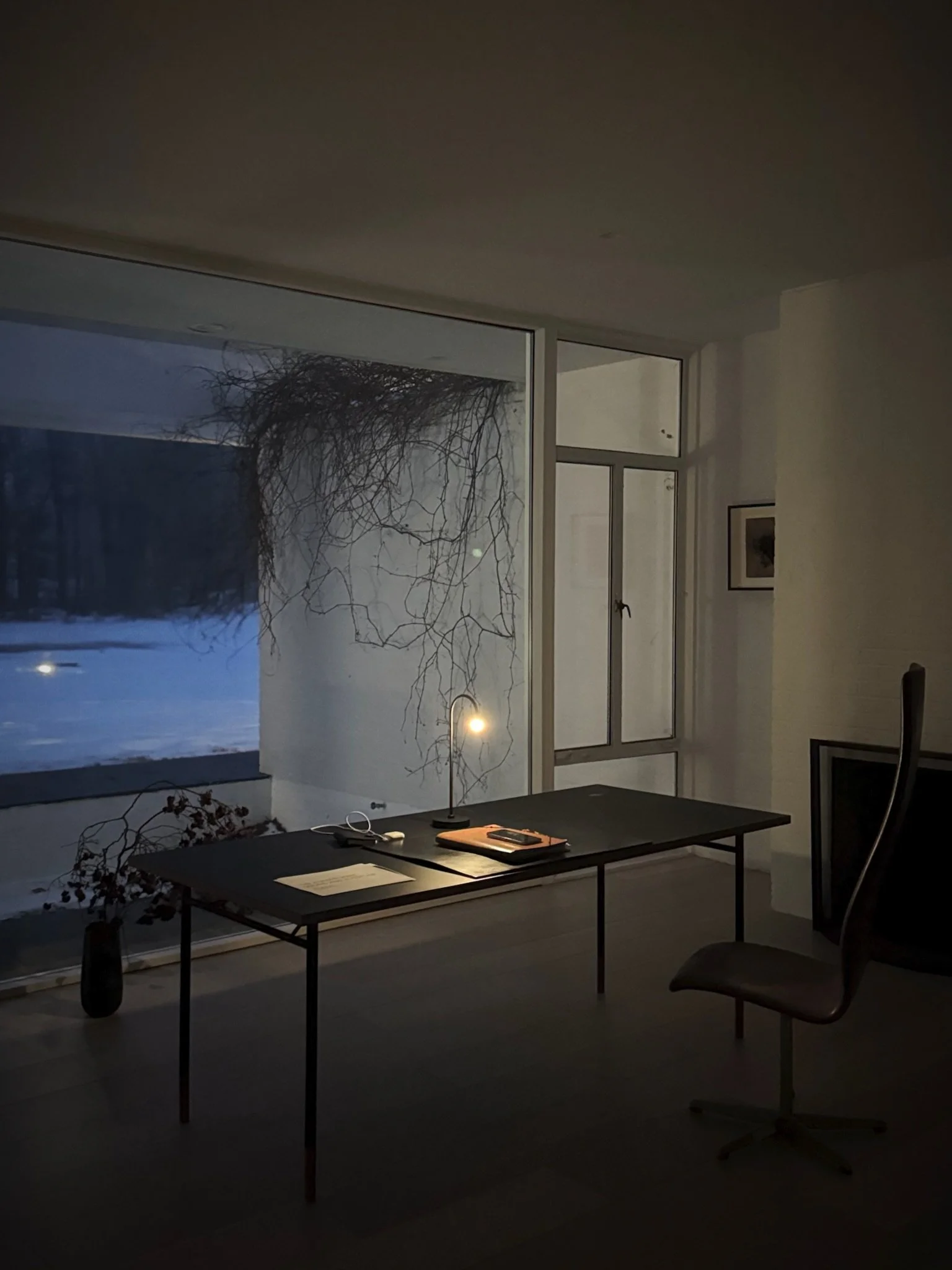 A minimalist office with a black desk, a chair, a desk lamp, and a view through large windows of a snowy landscape and leafless tree branches at dusk.