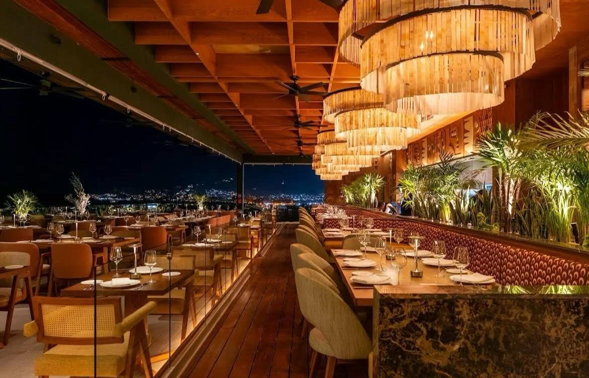 Interior de un restaurante elegante con mesas listas, iluminación cálida, plantas decorativas y vistas a la ciudad de noche.