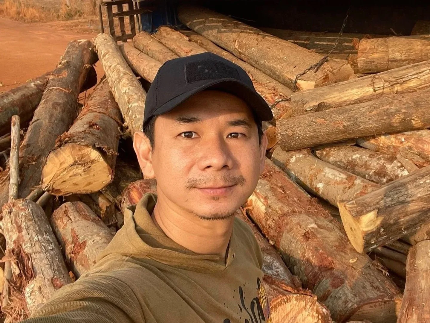 A man taking a selfie in front of a large pile of cut logs. The man is wearing a black cap and a brown hoodie. The logs are stacked on a truck or trailer.