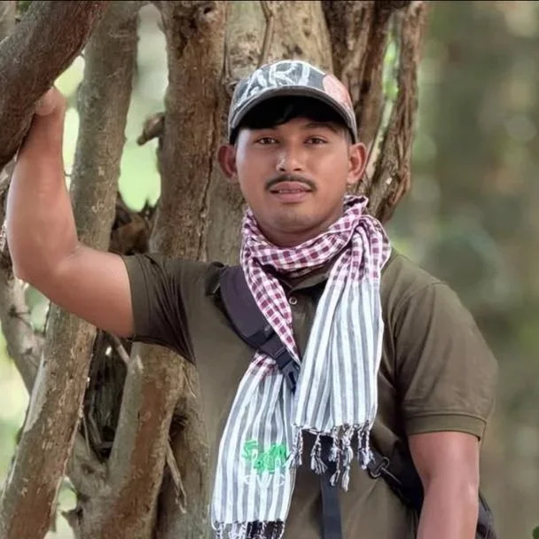 A man outdoors standing near a tree, wearing a baseball cap, a checkered scarf around his neck, a brown t-shirt, and carrying a backpack.