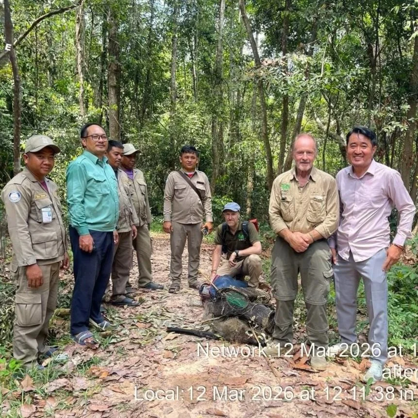 A group of seven men standing in a forest, some in khaki uniforms and others in casual attire, with a man crouching down behind some dead animals in a trap on the ground.
