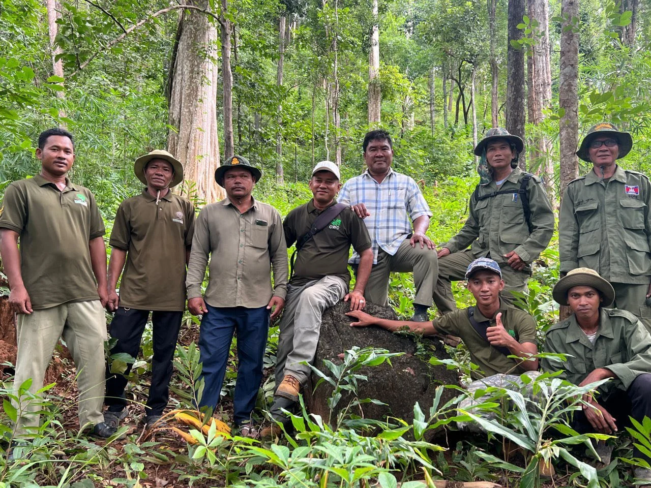 A group of ten men, mostly dressed in outdoor or field clothing, are standing and sitting among lush green trees and foliage in a forest. Some are wearing hats, and they are posing for a photo with smiles, showing camaraderie and teamwork.