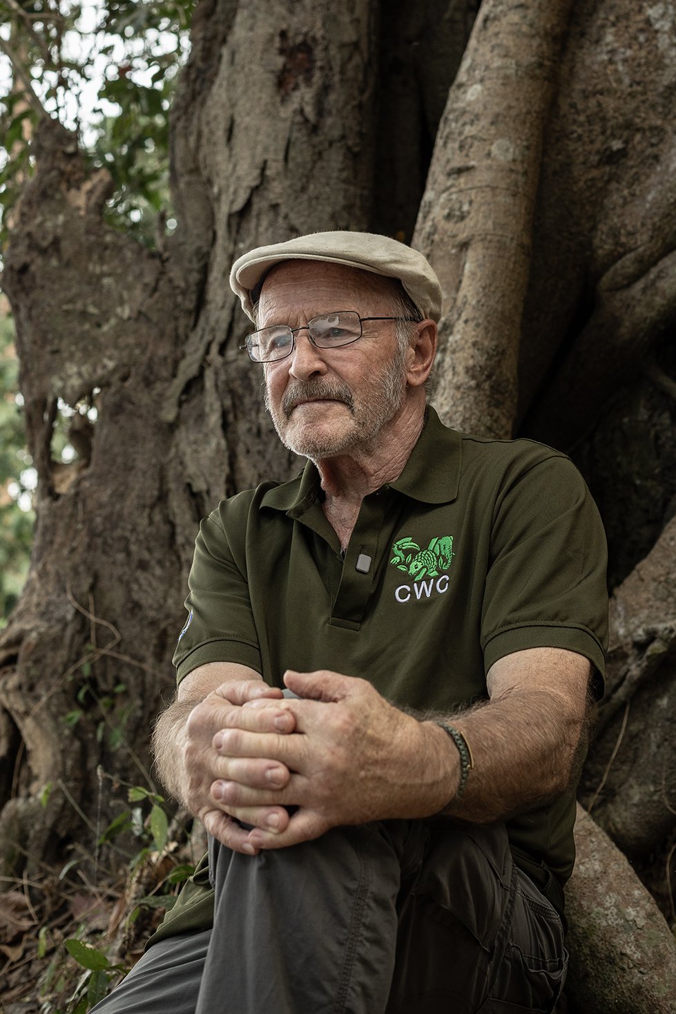 An elderly man wearing glasses, a beige cap, and a green polo shirt with a logo sitting against a large tree.