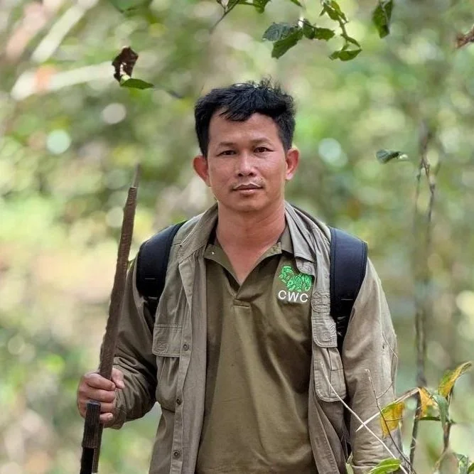 A man with short black hair outdoors, wearing a light jacket over a khaki shirt with a CWC logo, carrying a backpack and holding a walking stick, surrounded by greenery.