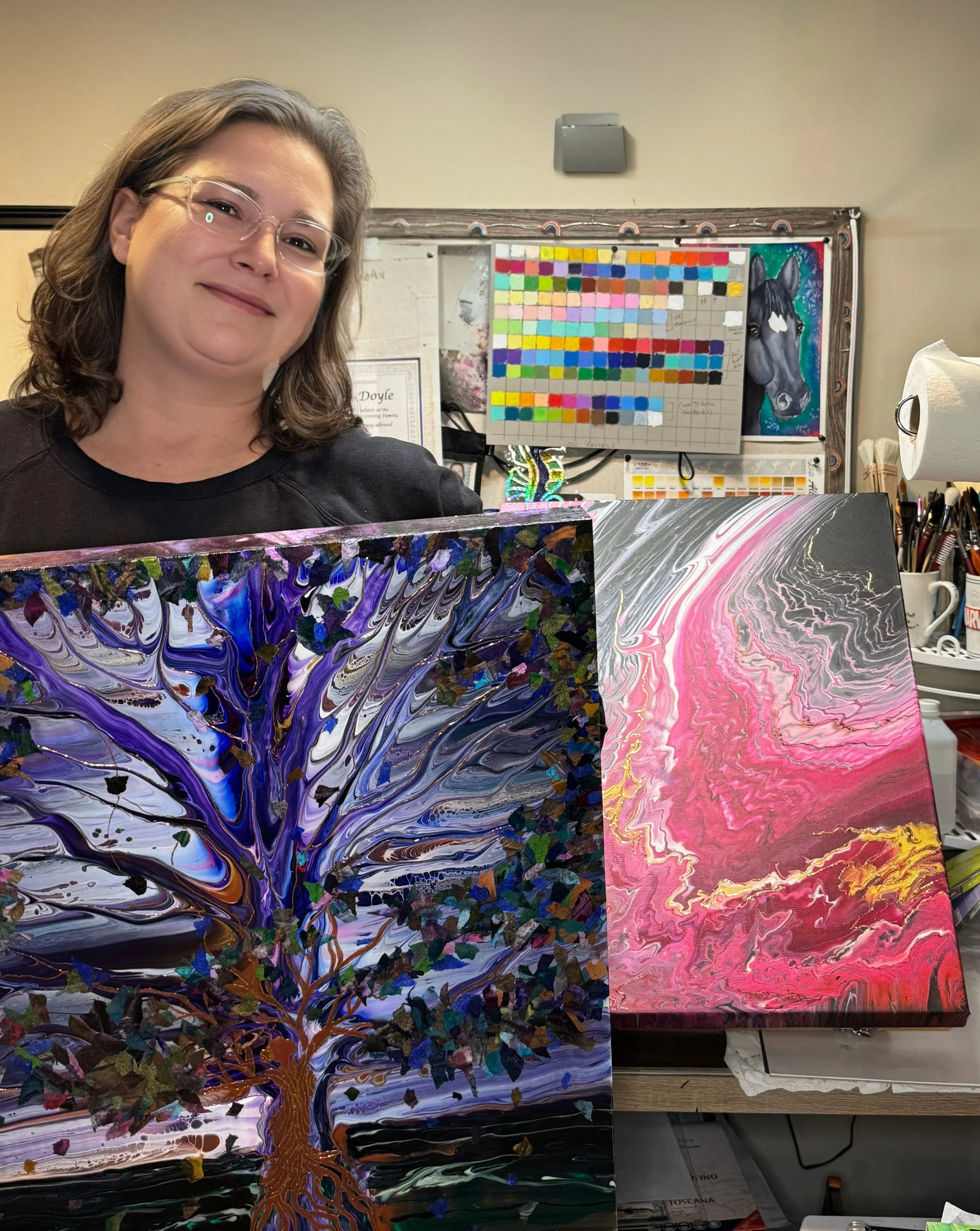 A woman with glasses and shoulder-length brown hair smiling behind two colorful abstract paintings in an art studio.