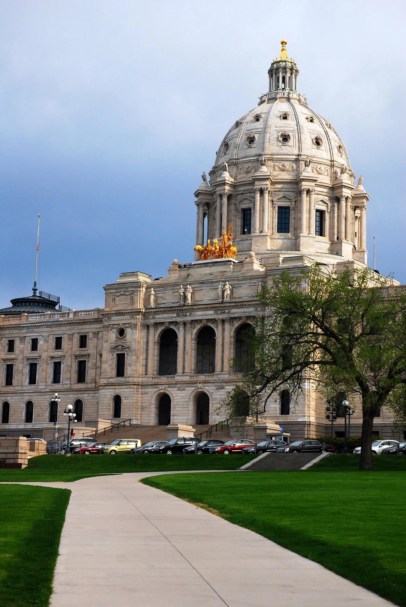 Photo of a historic government building with a grand domed roof and decorative sculptures, set against a cloudy sky. There are green lawn and trees in the foreground, with a parking lot containing several cars at the base of the building.