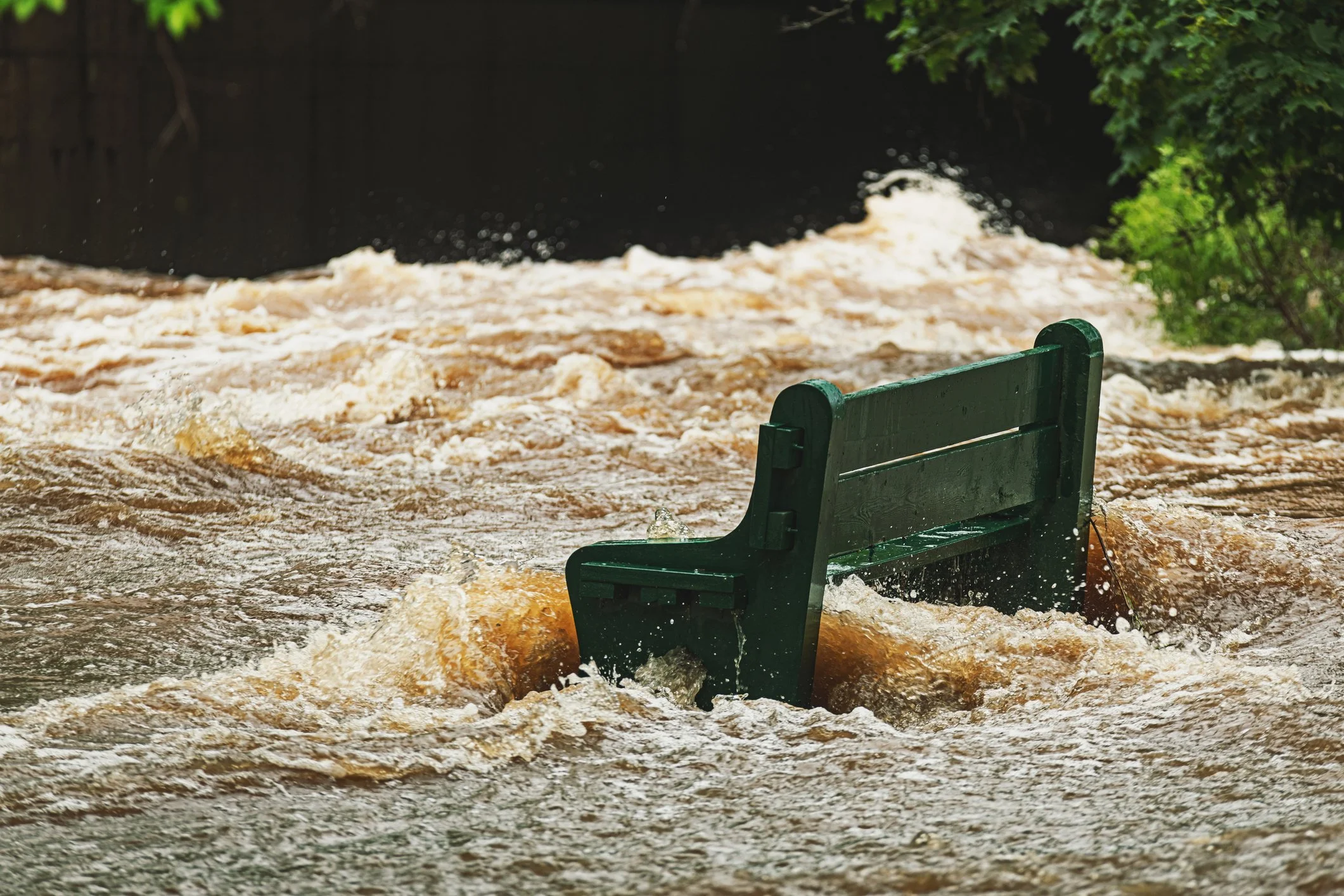 A park bench is partially submerged in floodwaters, with the water rushing around it and trees visible in the background.