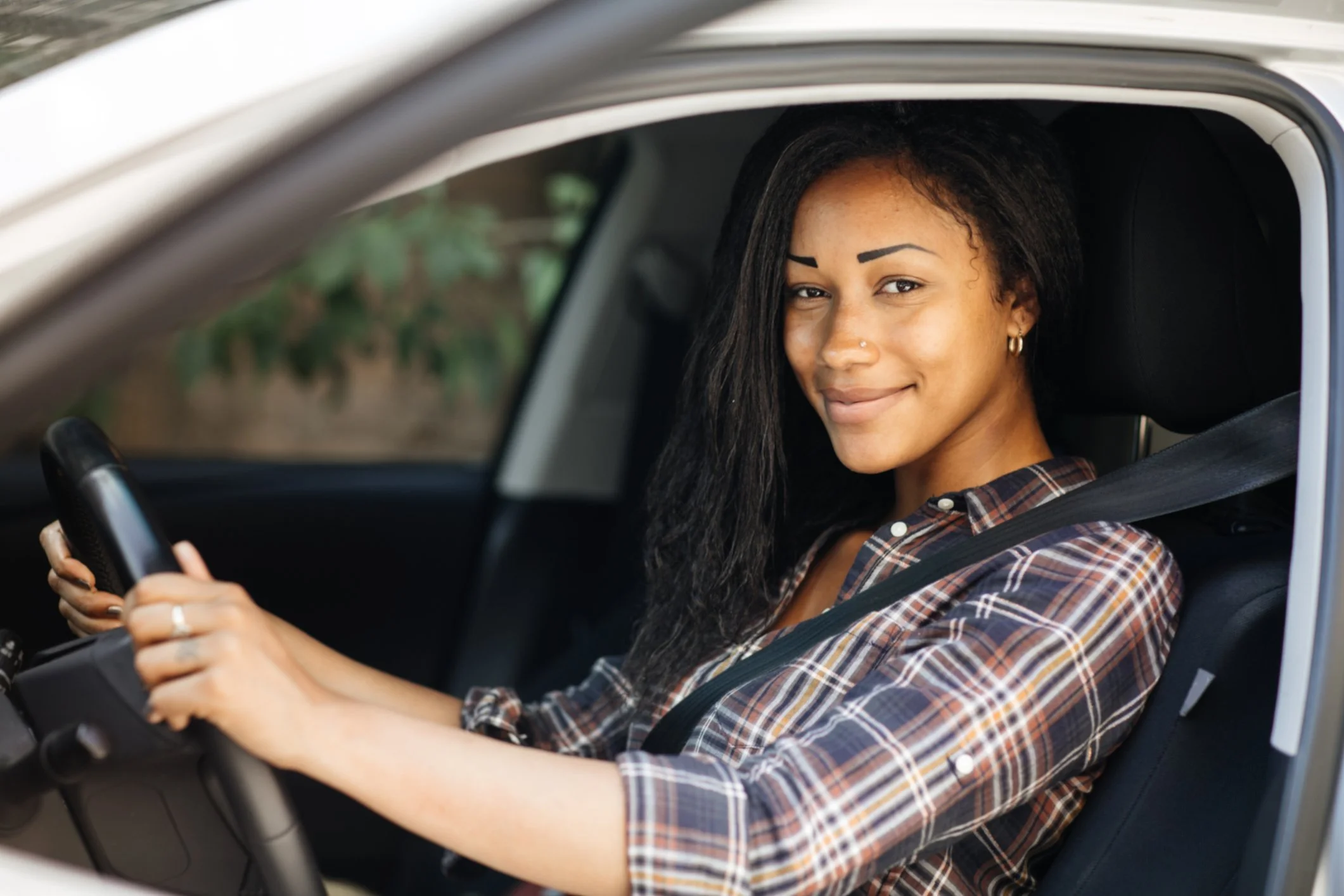 A woman wearing a plaid shirt and a seatbelt sitting in a car with her hands on the steering wheel, smiling at the camera.