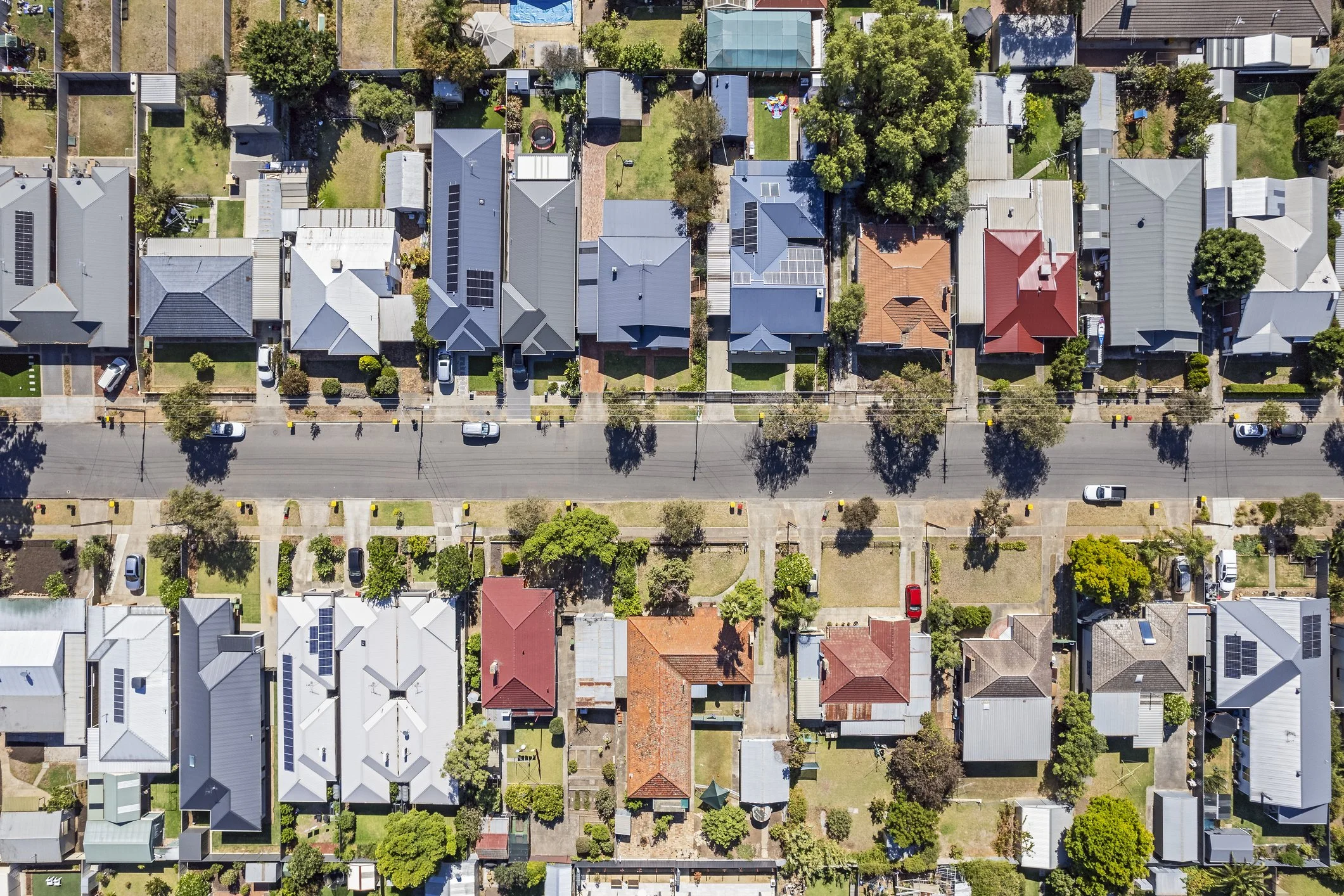 Aerial view of a residential neighborhood showing houses with various roof colors and styles, trees, yards, and a main street with parked cars.