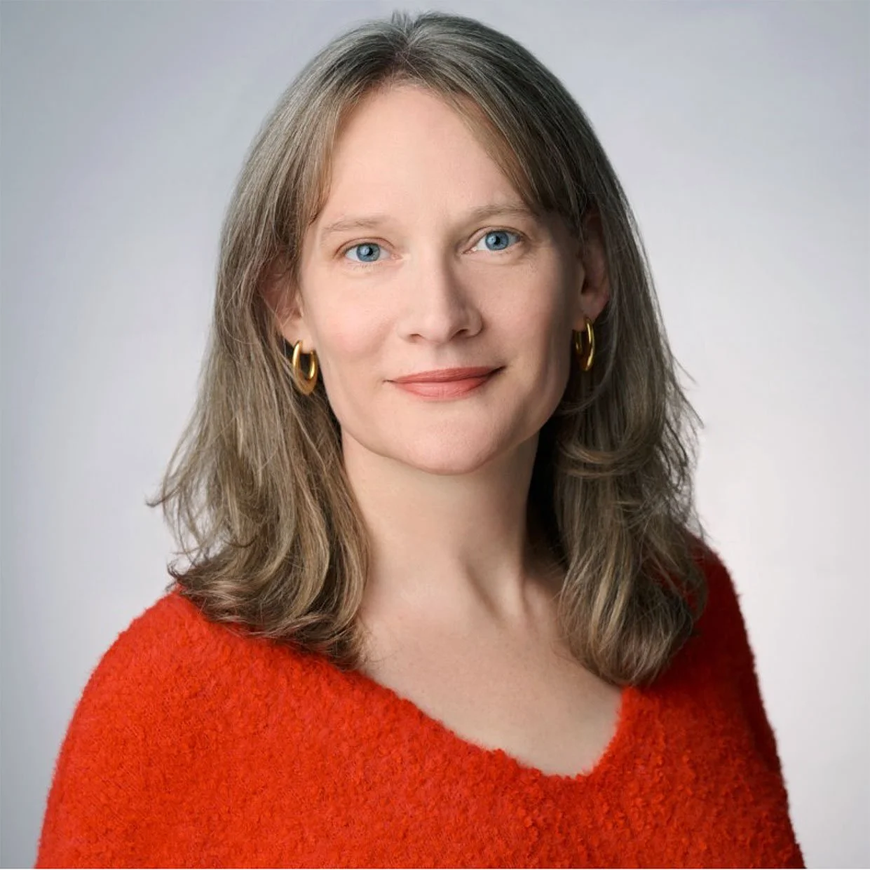 A woman with shoulder-length brown hair, blue eyes, wearing gold hoop earrings and a red textured sweater, smiling against a light gray background.