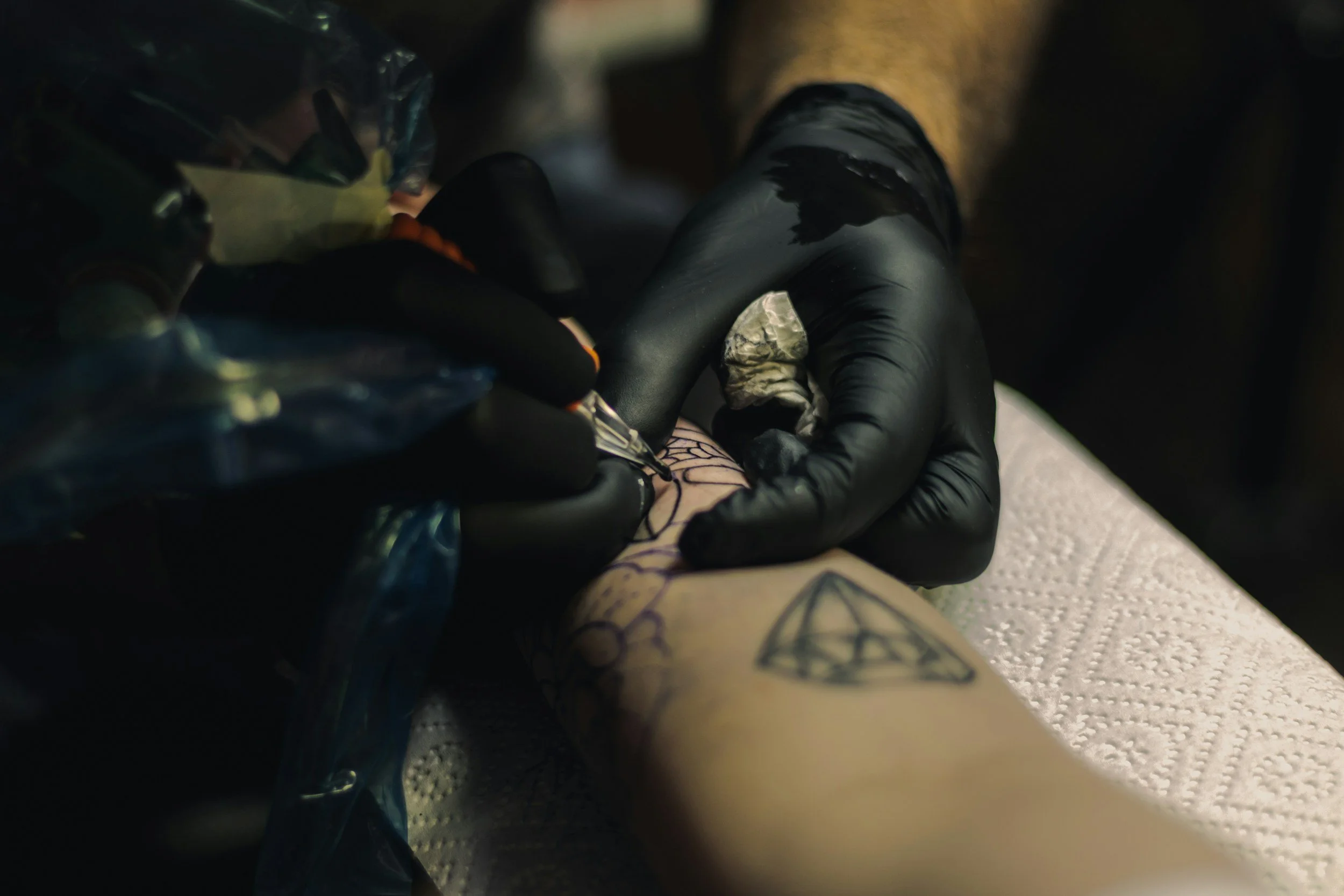 A person wearing black gloves is tattooing a geometric design on a person's forearm, with the tattoo partially filled in, on a white paper towel.