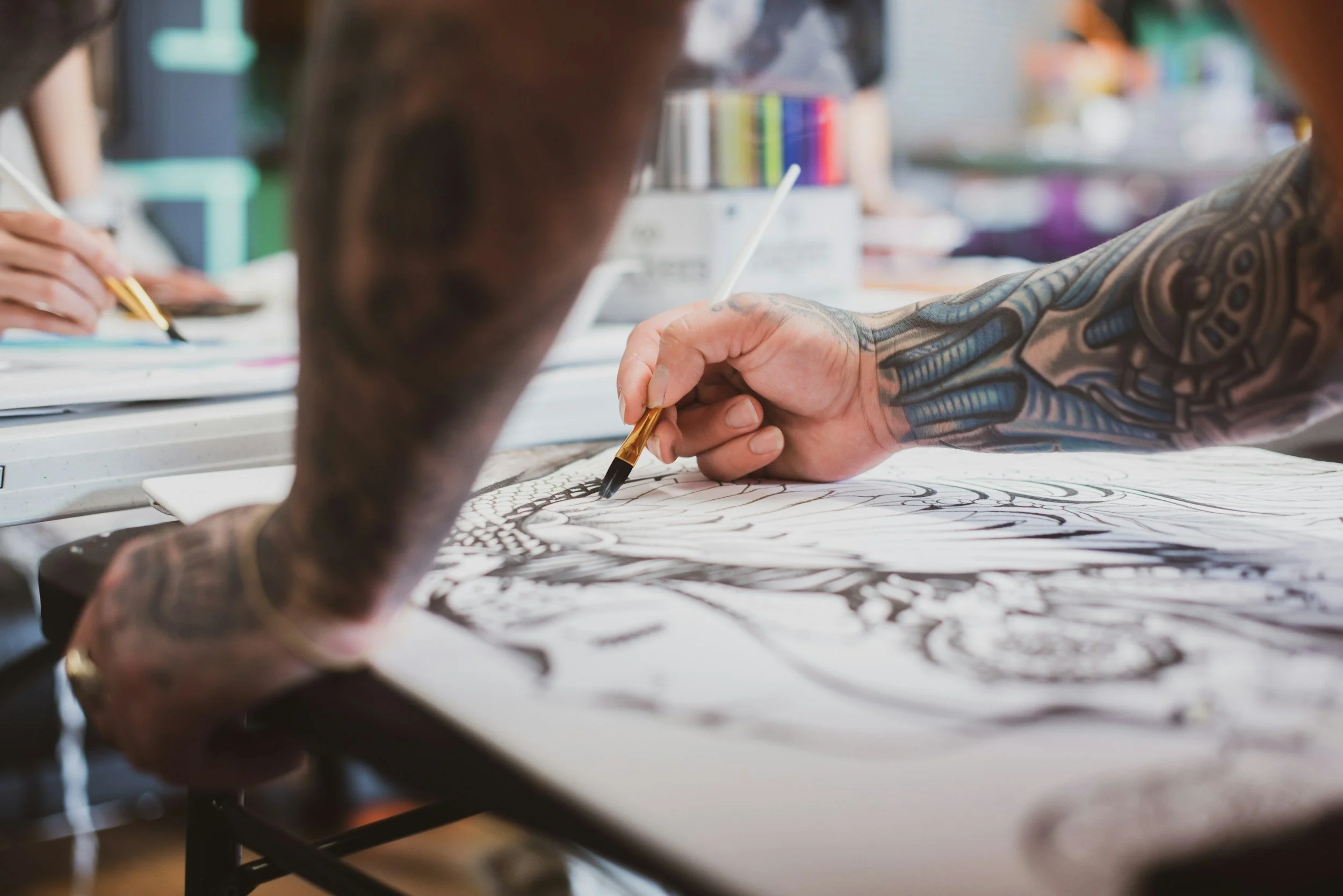 Tattooed person drawing on large sheet of paper with black ink, surrounded by art supplies in a bright studio.