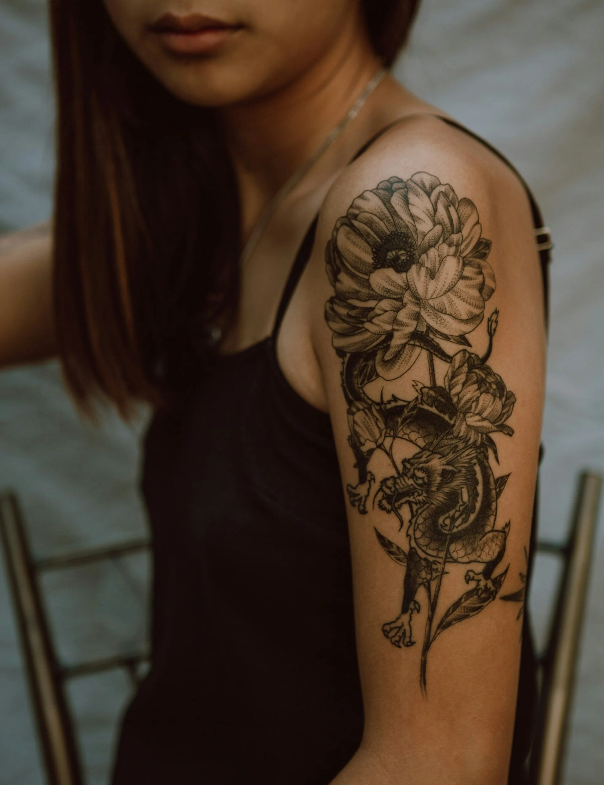 A woman with long brown hair and a black sleeveless top with spaghetti straps, showing a large black and gray tattoo of a dragon intertwined with flowers on her upper arm and shoulder.