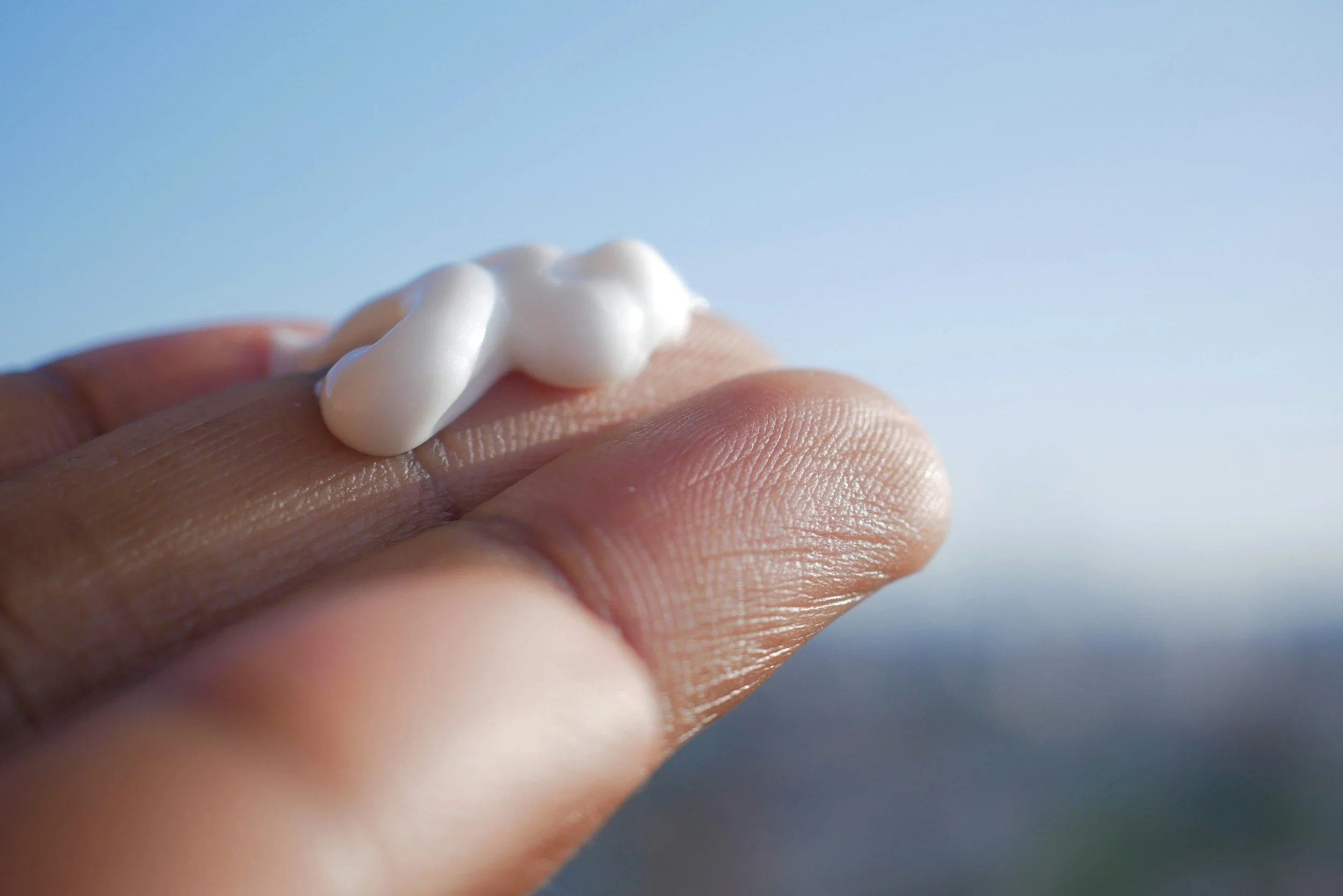 Close-up of a person's finger with a dollop of white cream on it, with a blurred blue sky background.